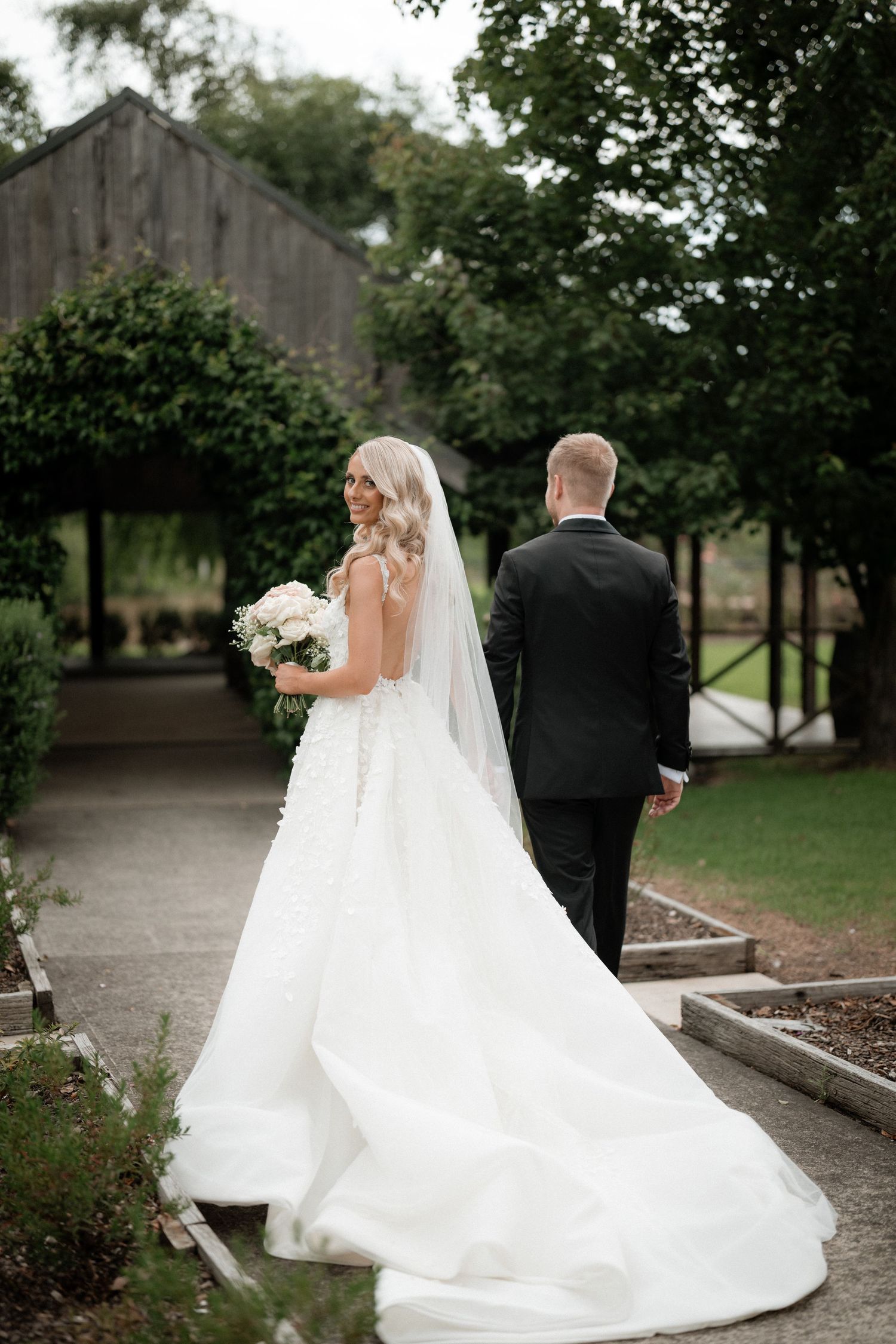 A bride in a flowing white wedding dress and groom walking outdoors near a rustic barn.