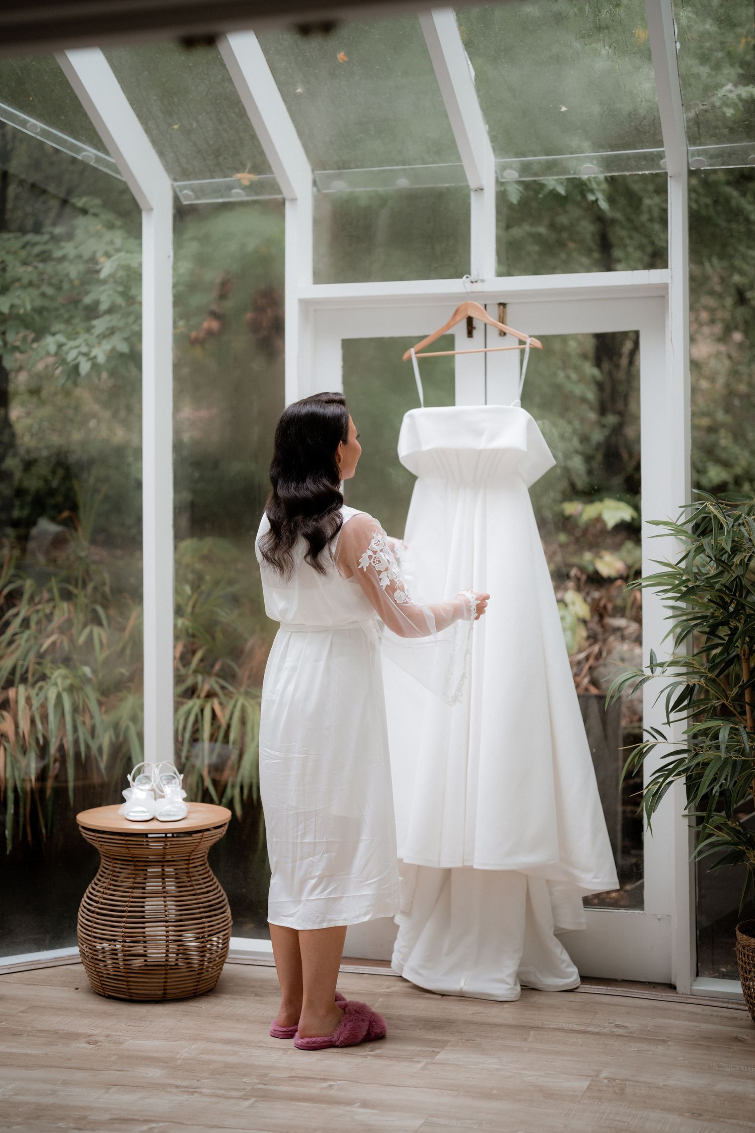A white wedding dress hangs in a sunlit conservatory while someone in a white robe reaches up to touch it.