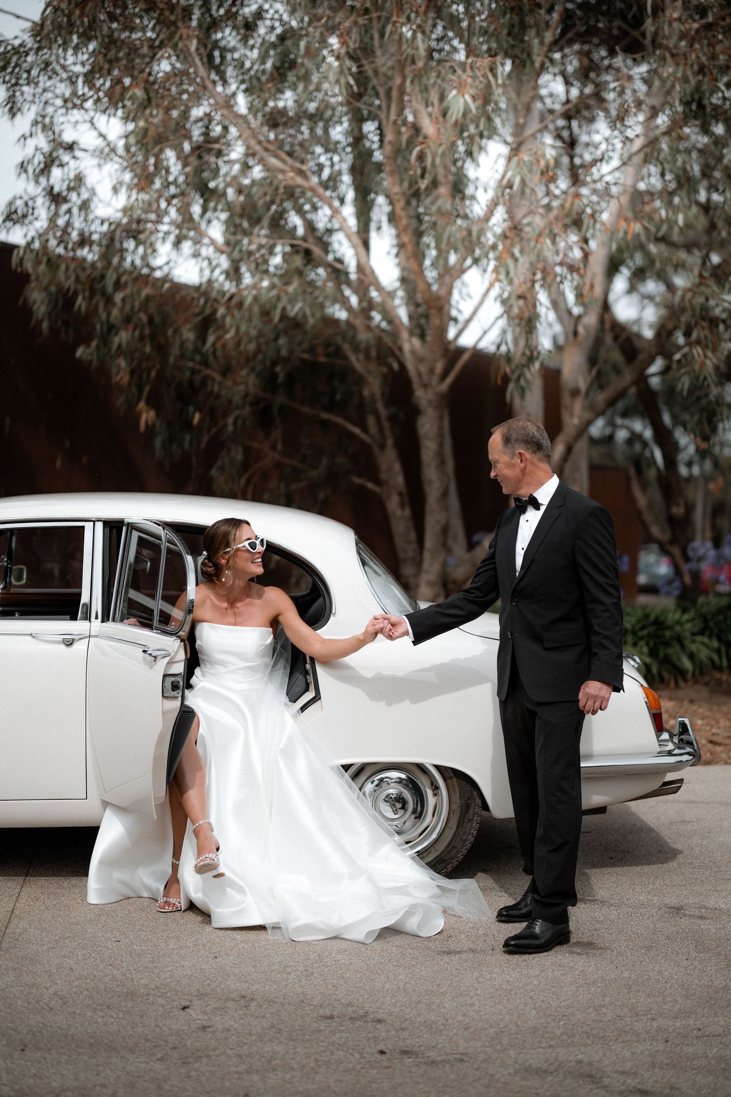 A couple in wedding attire poses beside a classic white vintage car under eucalyptus trees.