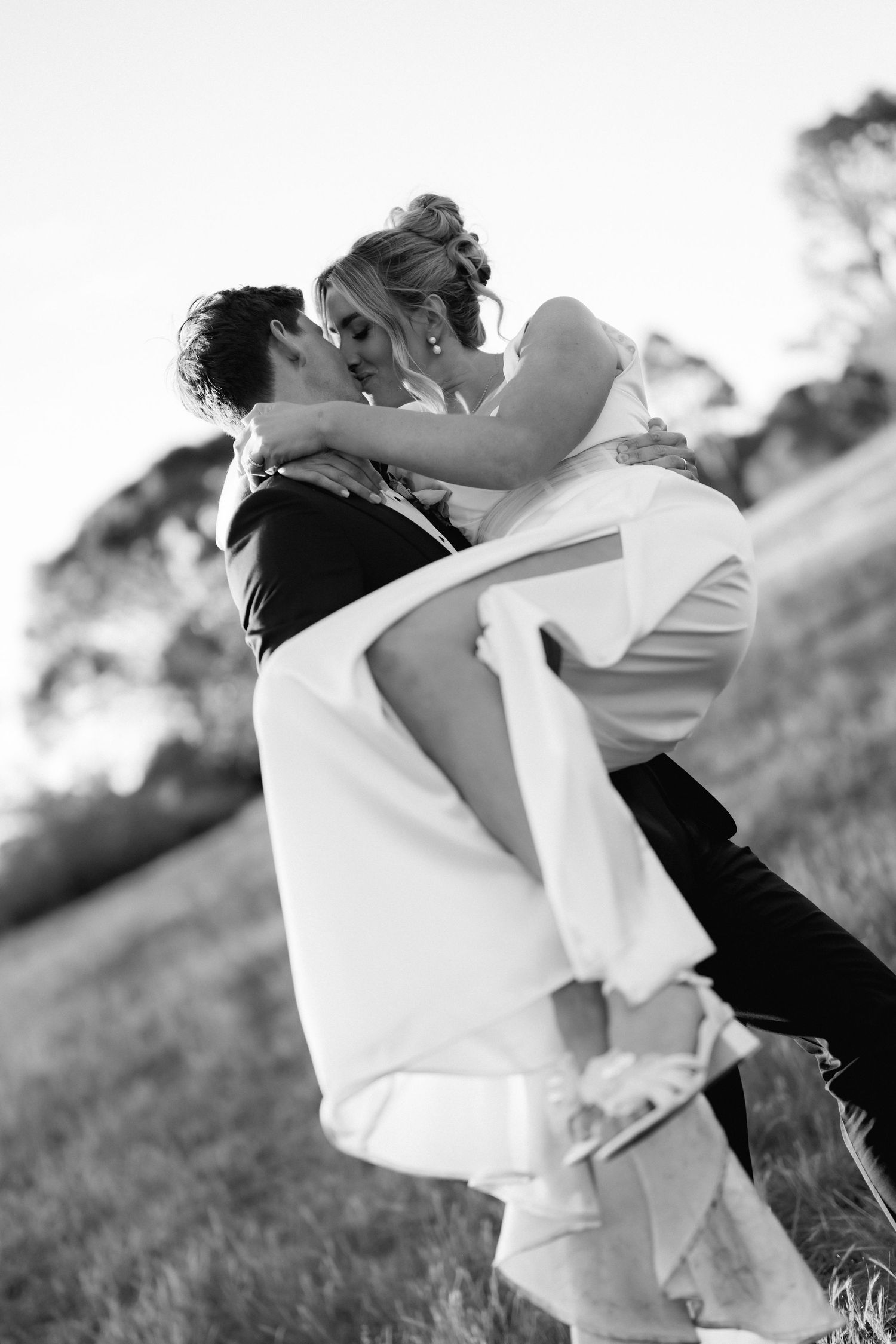 A black and white photo captures a romantic dip pose between newlyweds on a grassy hillside.