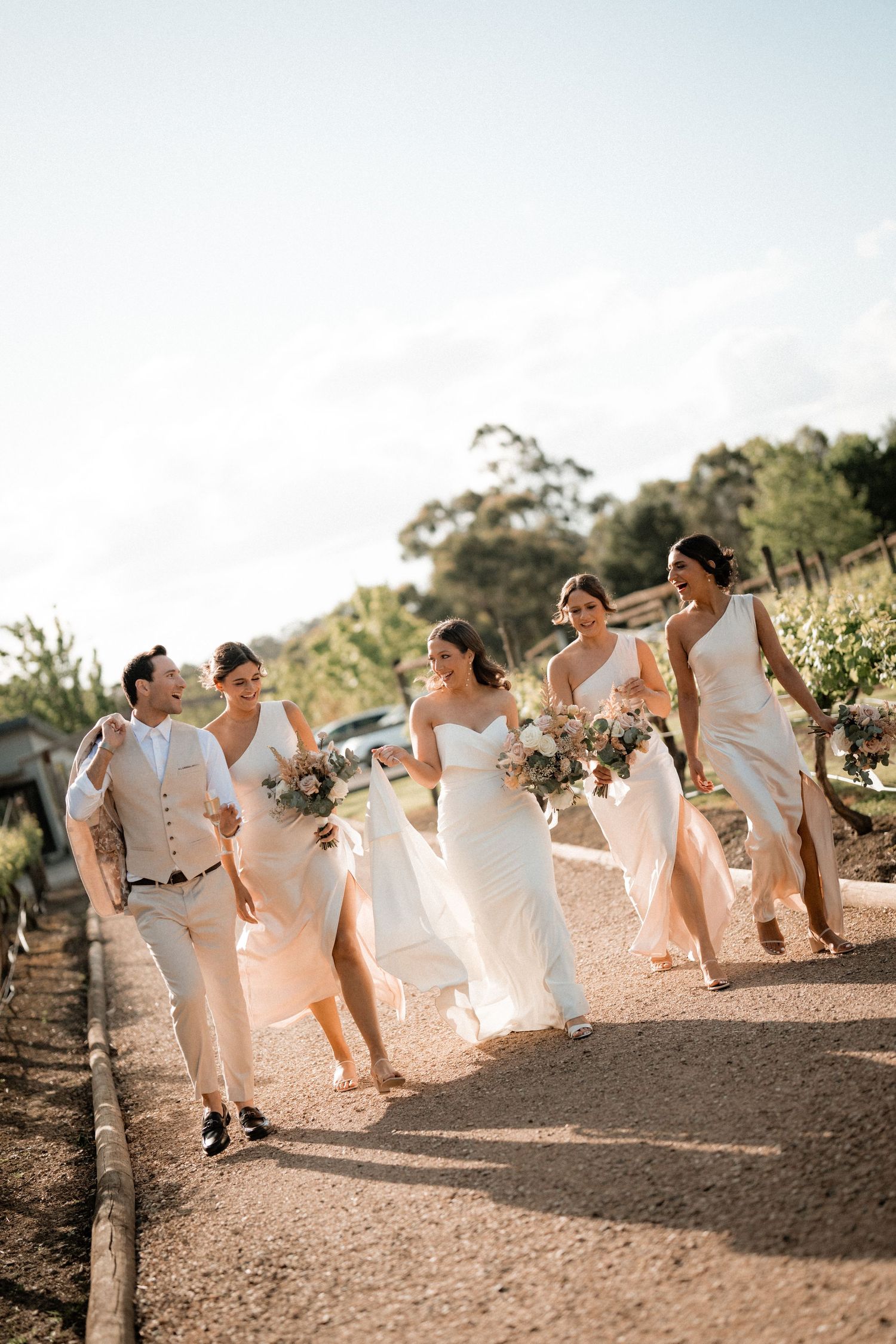 Wedding party jumps in coordinated white and beige attire against a golden sunset backdrop on a dirt path.
