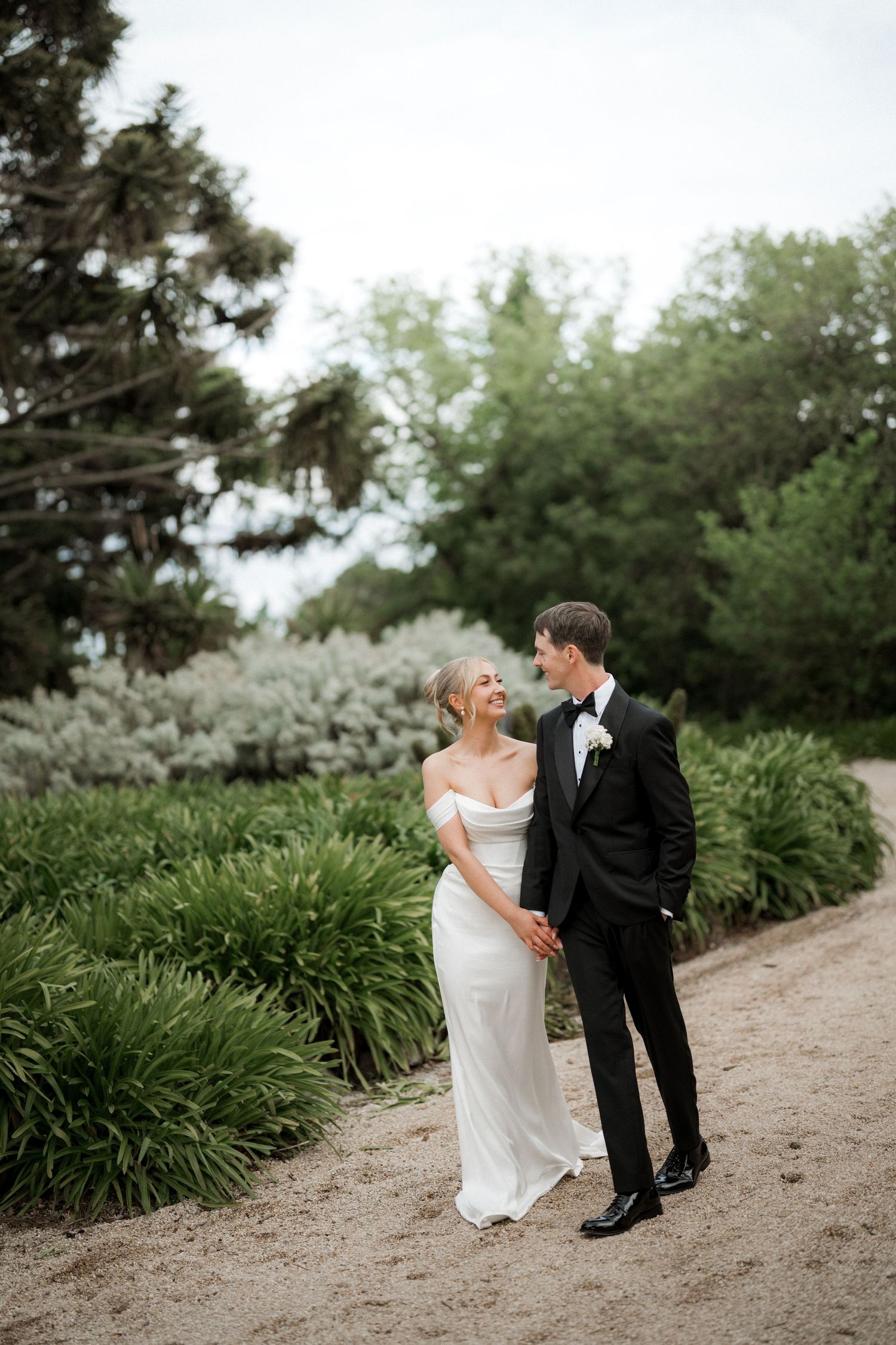 A couple shares a romantic moment on a garden path lined with lush greenery.
