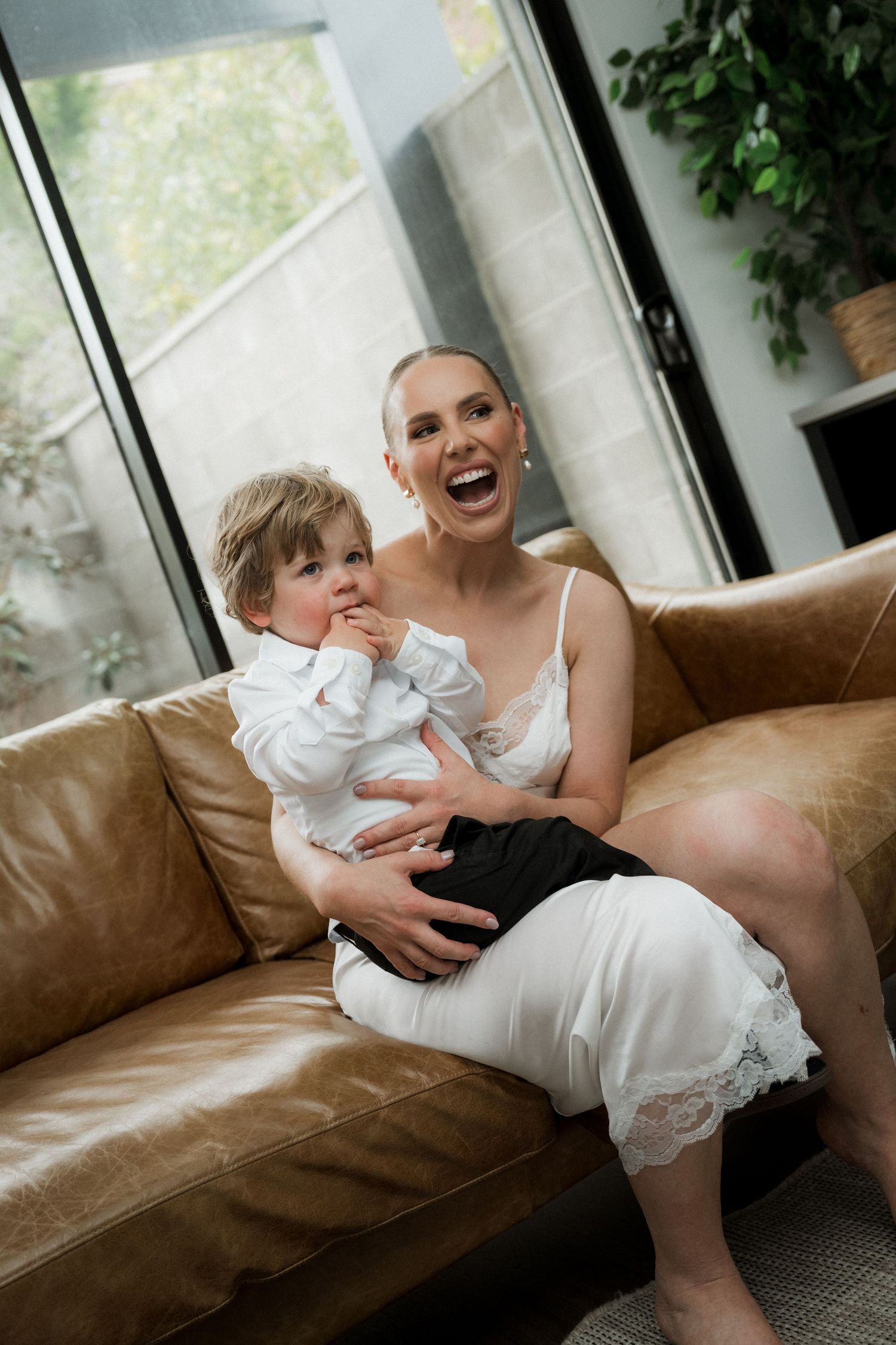 Joyful moments captured on a brown leather couch in a sunlit room with white wedding attire.