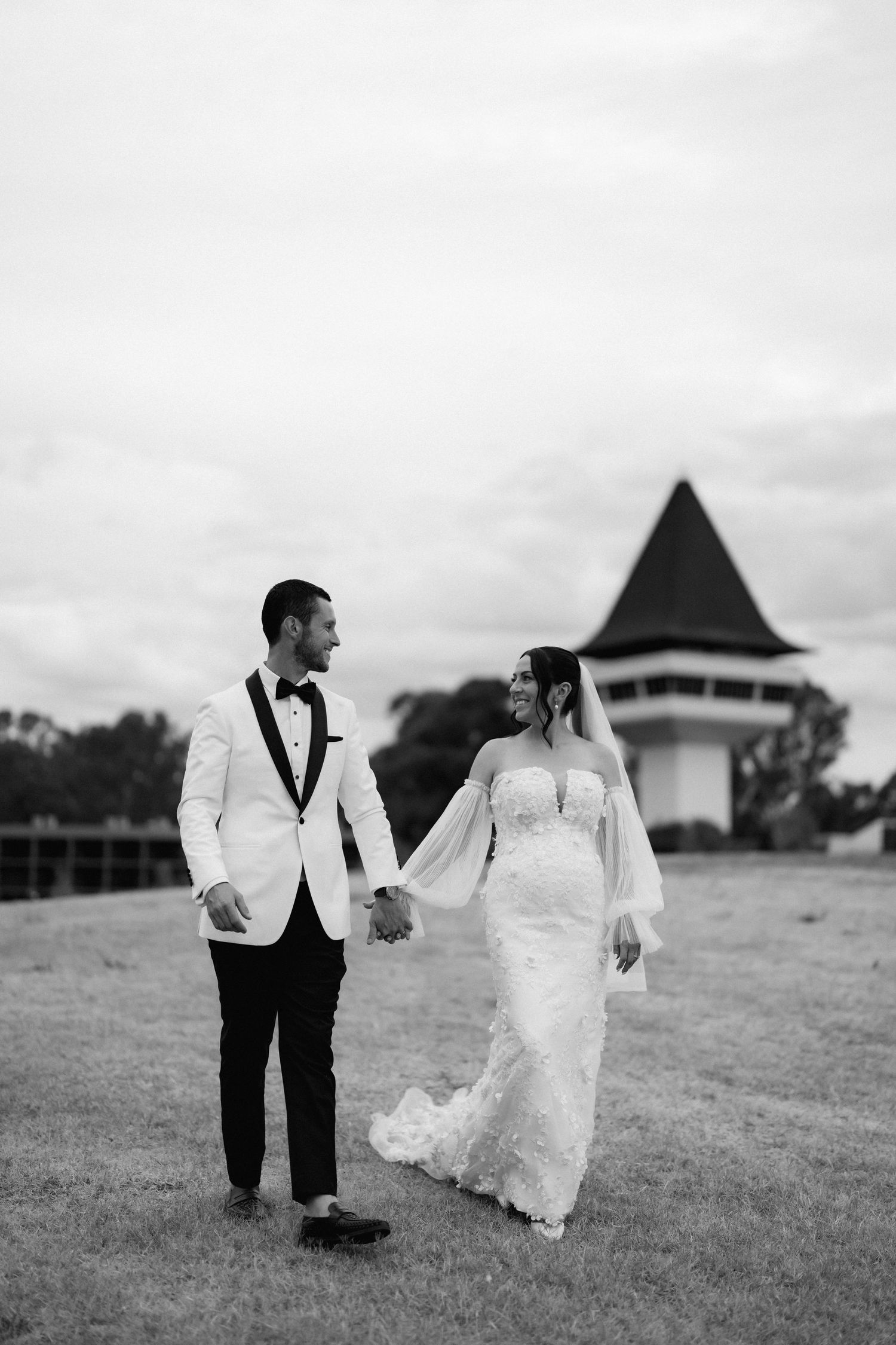 A couple in wedding attire dances playfully on a grassy lawn with a building in the background.