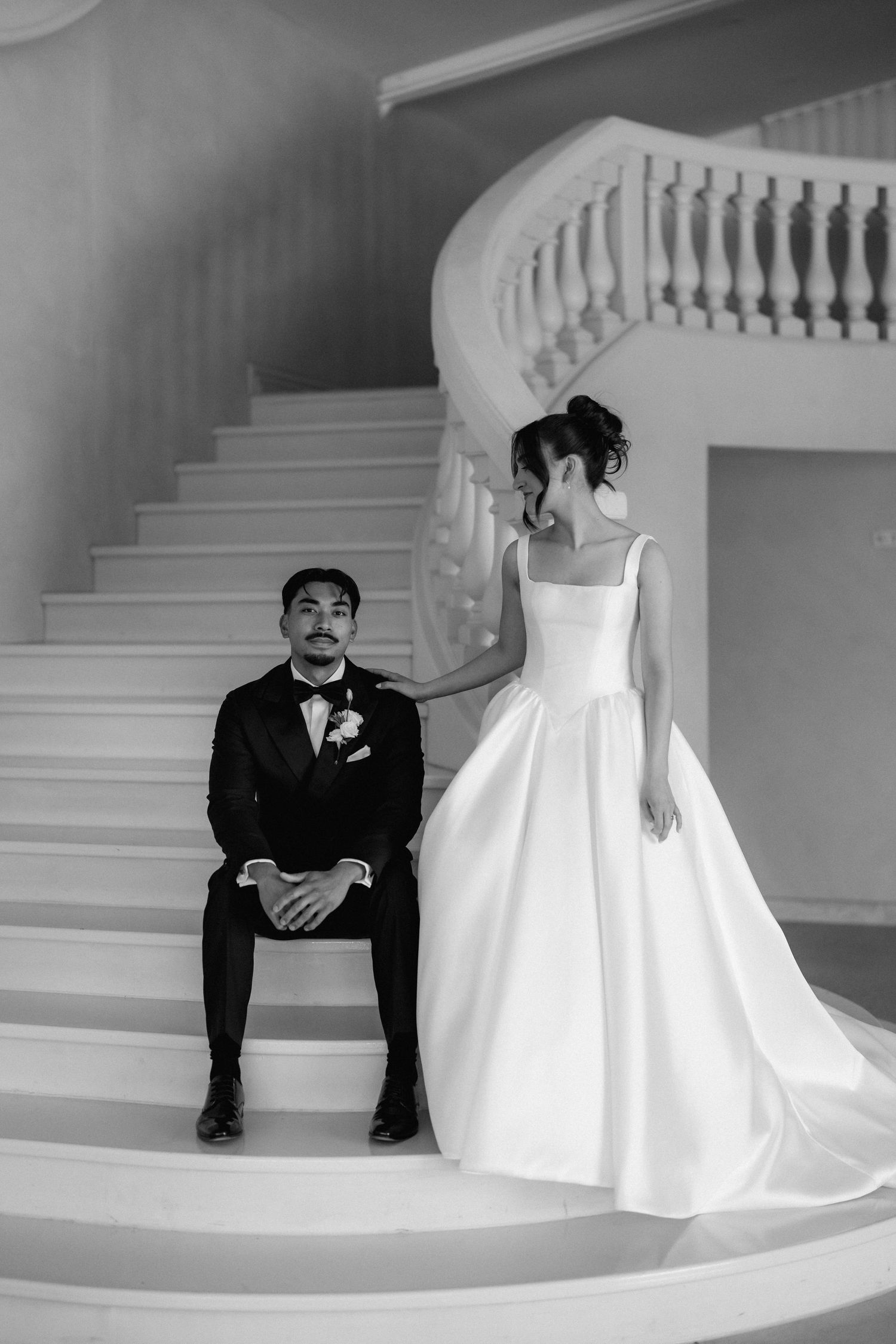 Black and white photo of a groom in a tuxedo seated on grand curved stairs as the bride in a satin ball gown stands beside him.