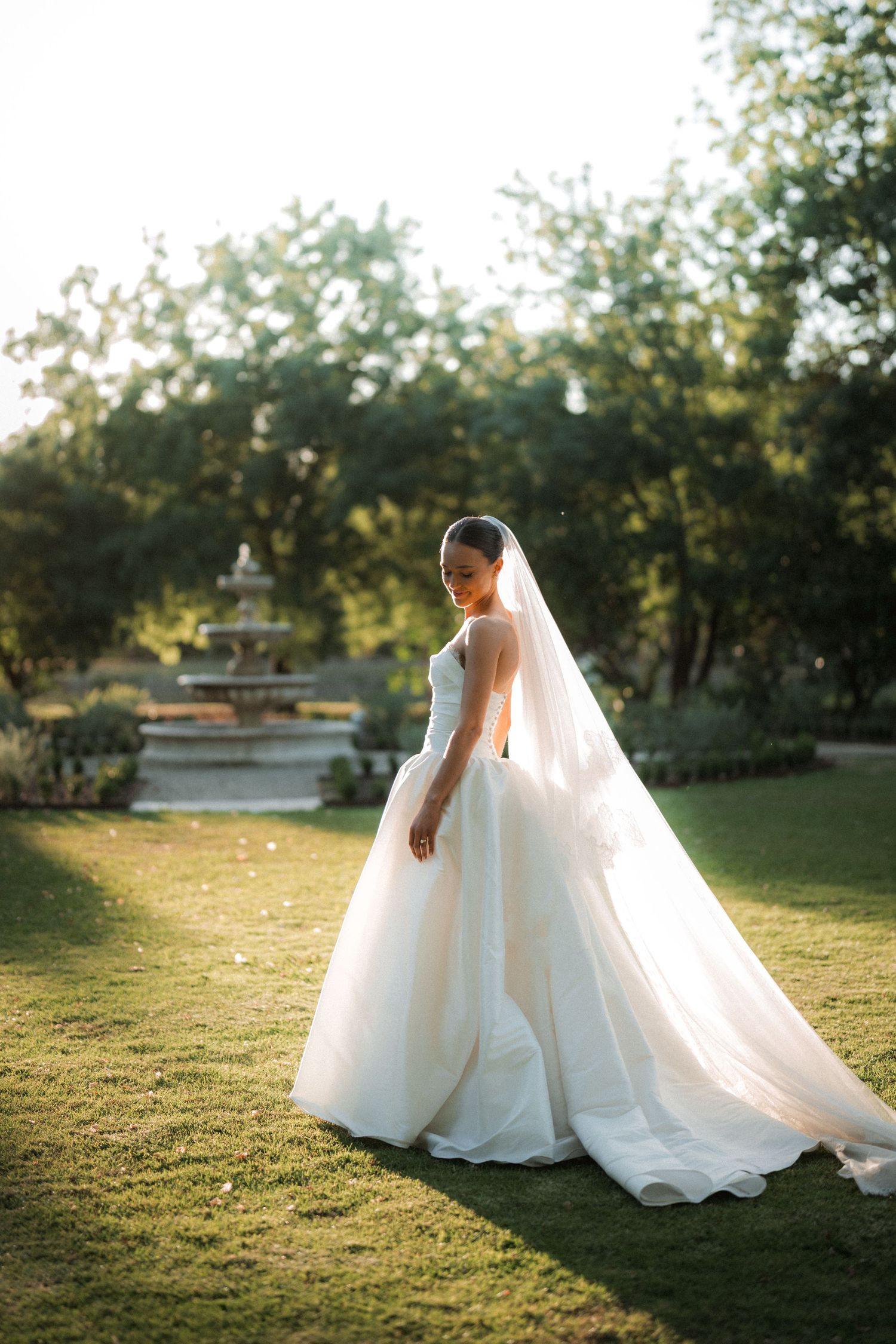 A bride in a flowing white gown walks gracefully through a sunlit garden setting.