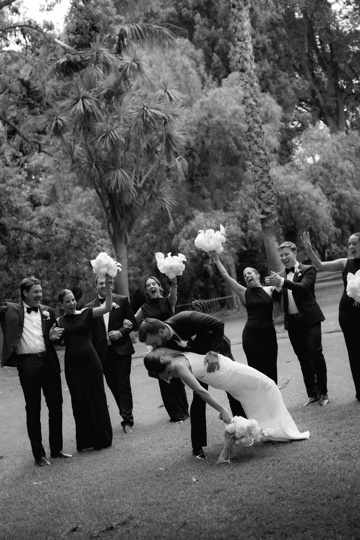 Black and white photo of wedding party celebrating with bride and groom dipping kiss.