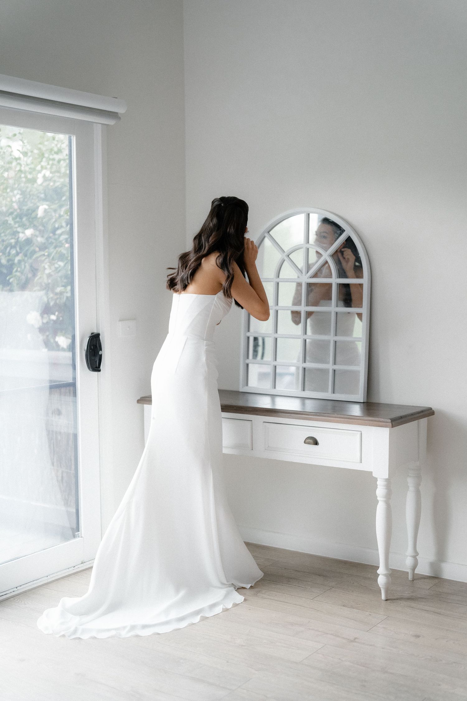 Bride in strapless white gown adjusting hair at vintage white console table with arched mirror.