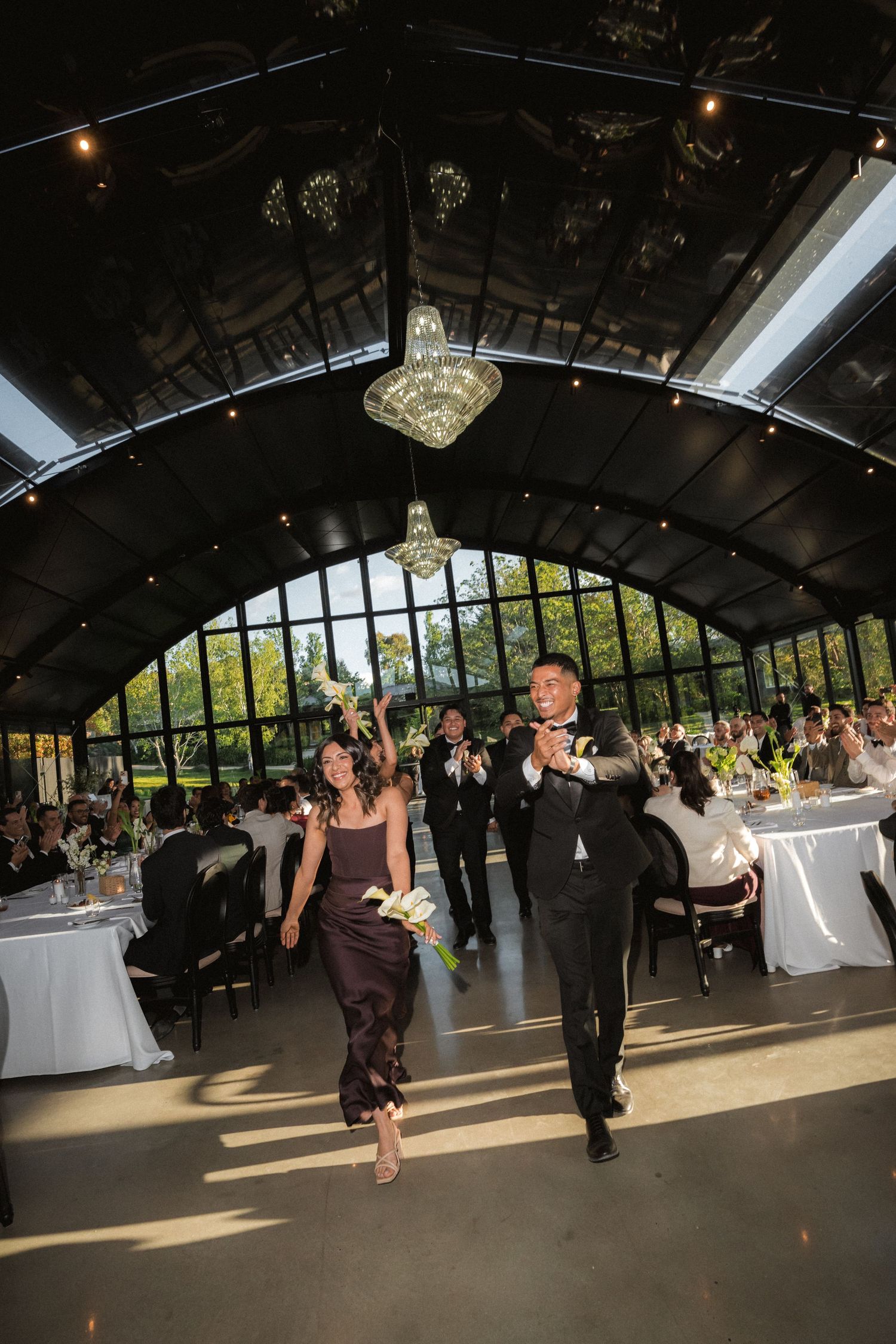 Bridesmaid in a plum dress and groomsman in a tuxedo walk in a wedding procession beneath chandeliers in a glass-walled reception hall.