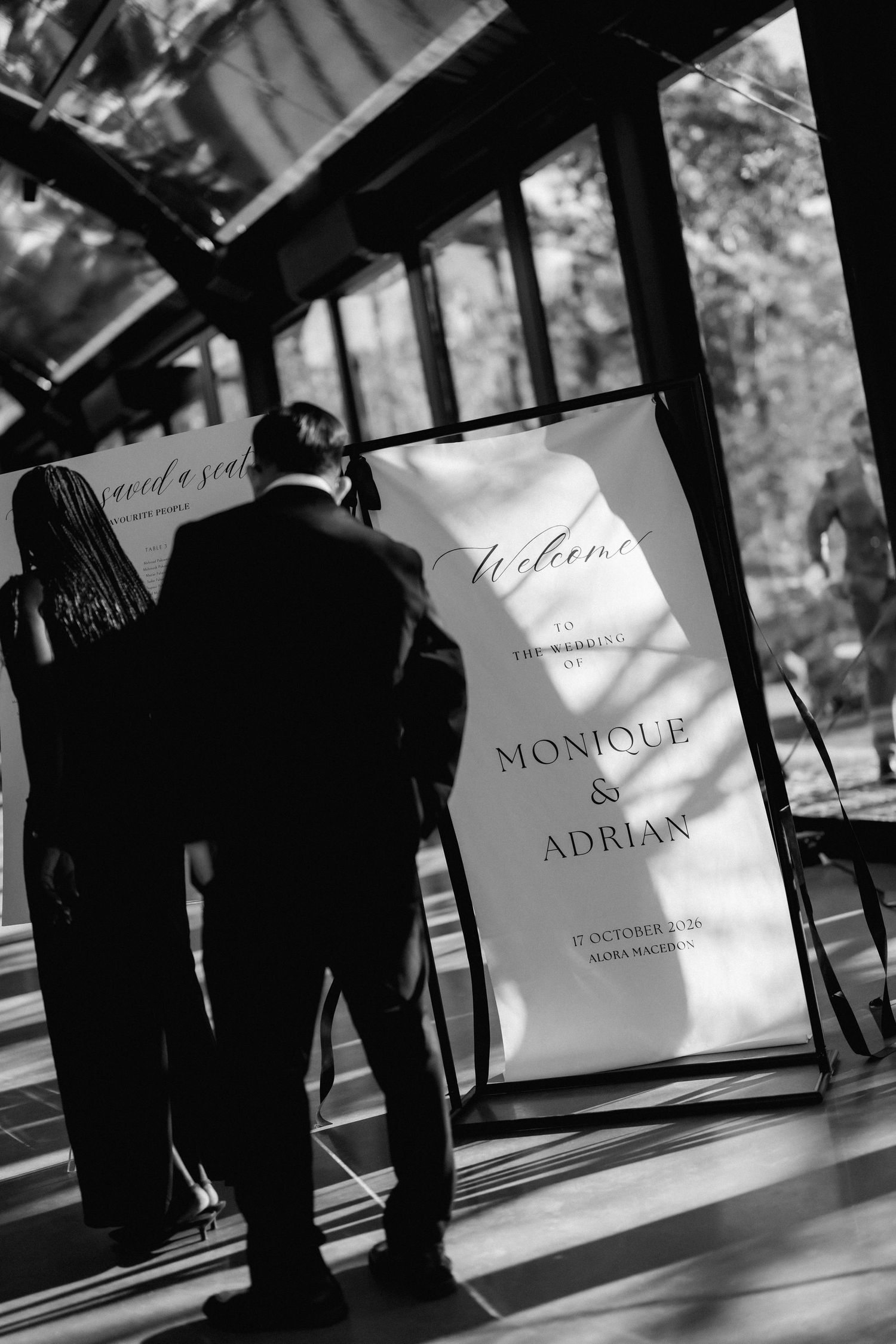 Black and white photo of wedding guests reading a welcome sign for Monique and Adrian's wedding inside a sunlit glass venue.