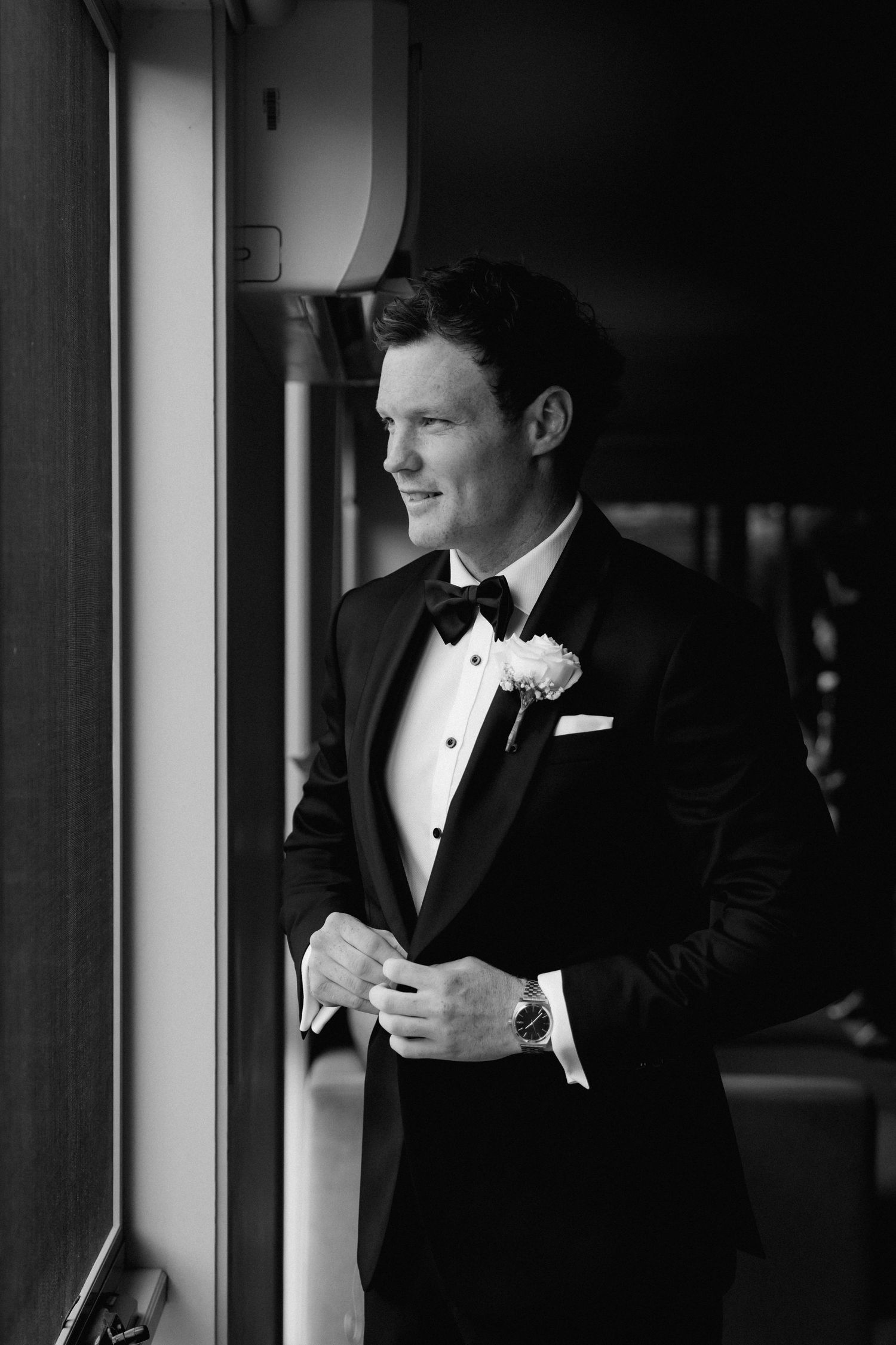 A groom in a classic black tuxedo and bow tie stands by a window in an elegant indoor setting.