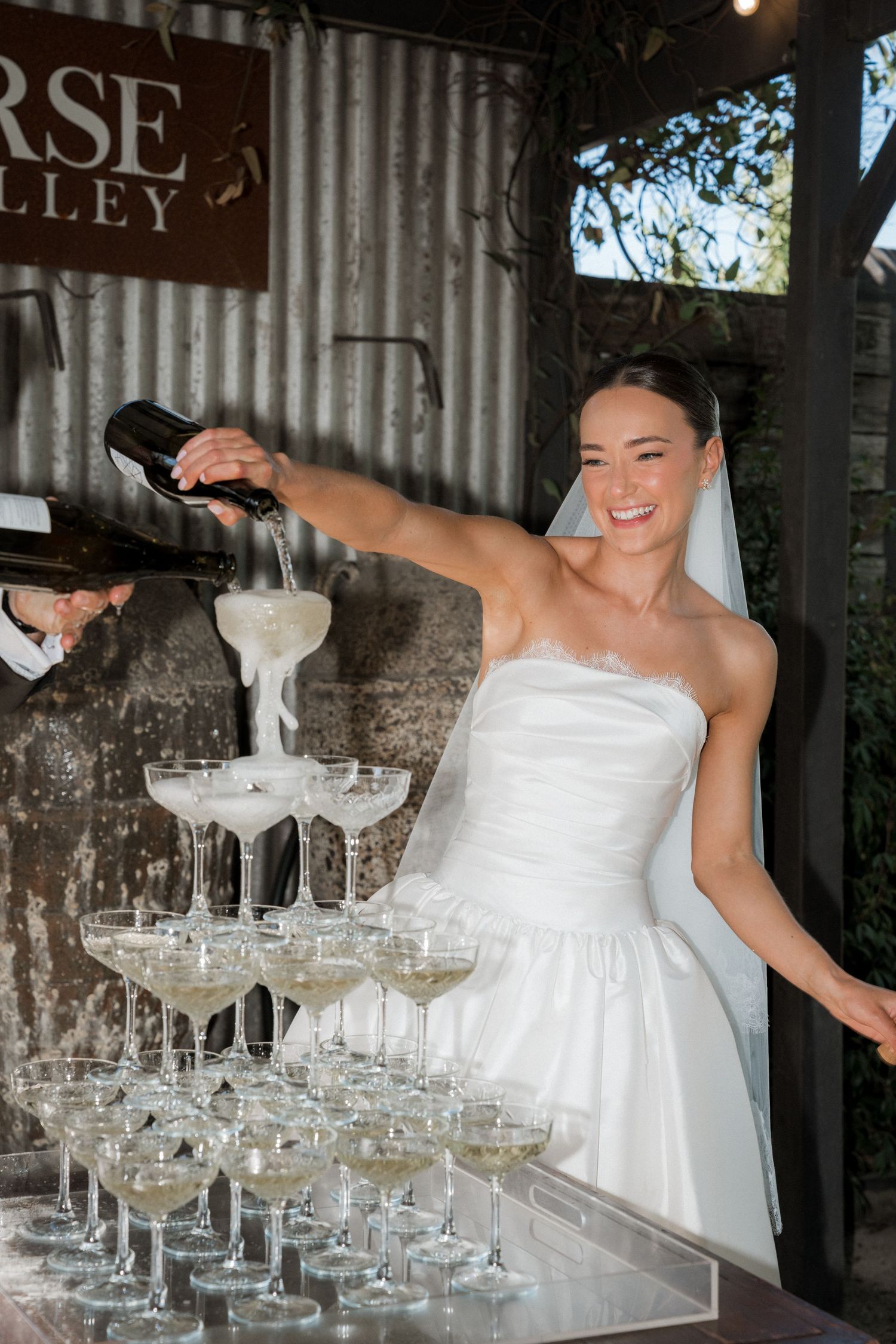 A bride in a white gown stands near a champagne tower display at a wedding reception.