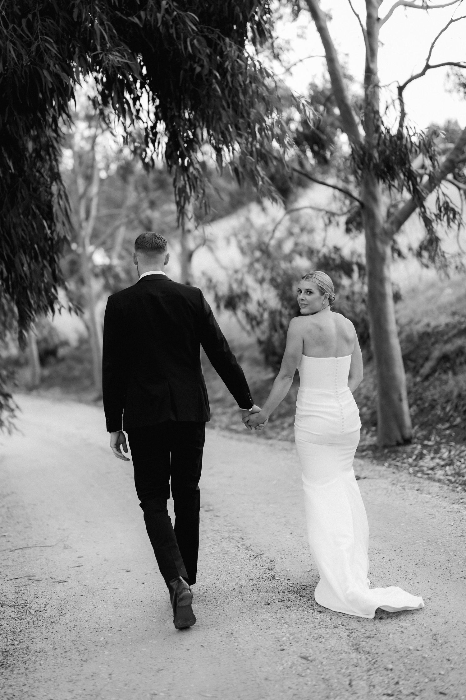Newlyweds walking hand in hand down a tree-lined path in black and white.