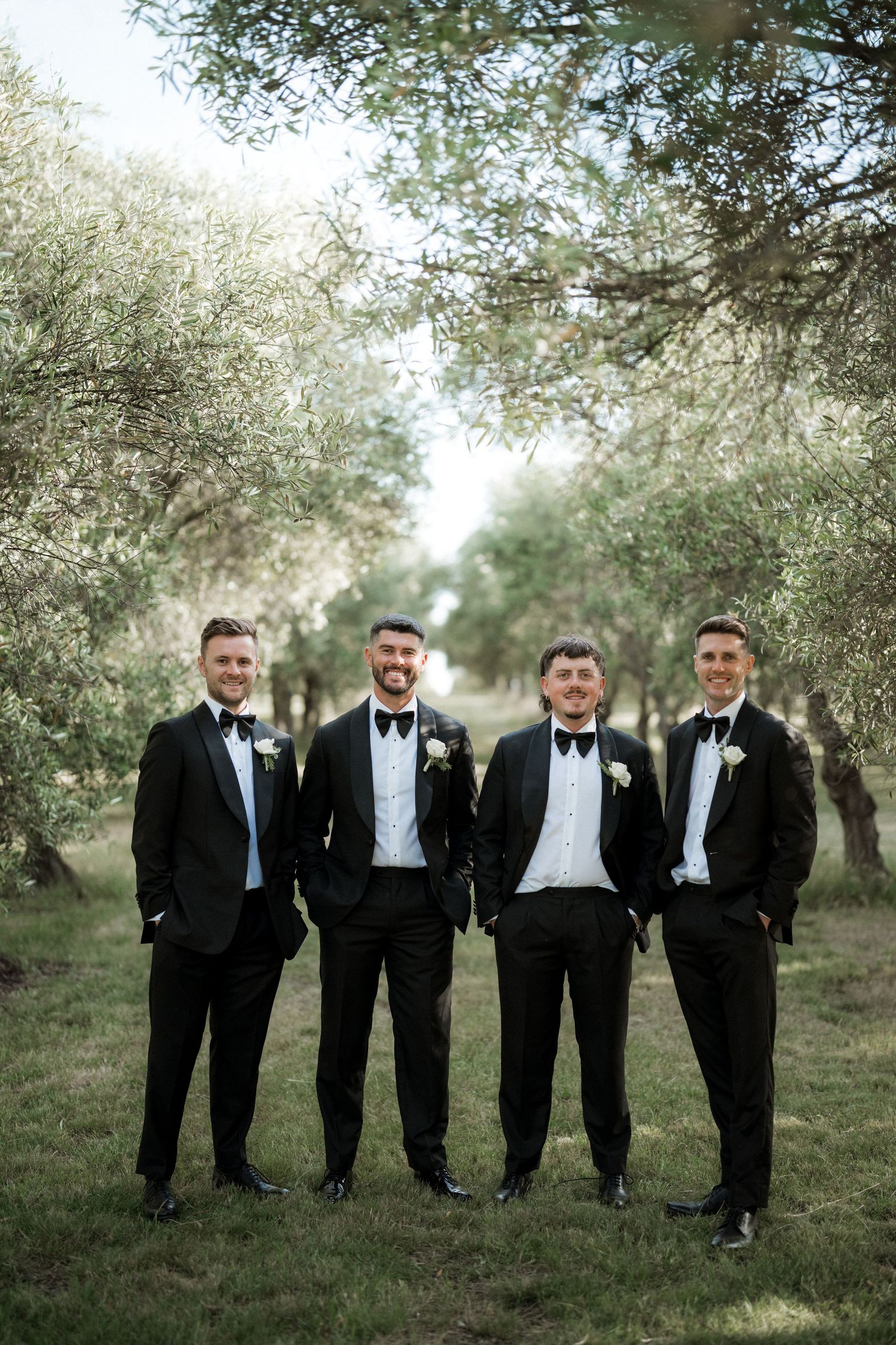 Groomsmen in black tuxedos posing together under trees for formal wedding photos.