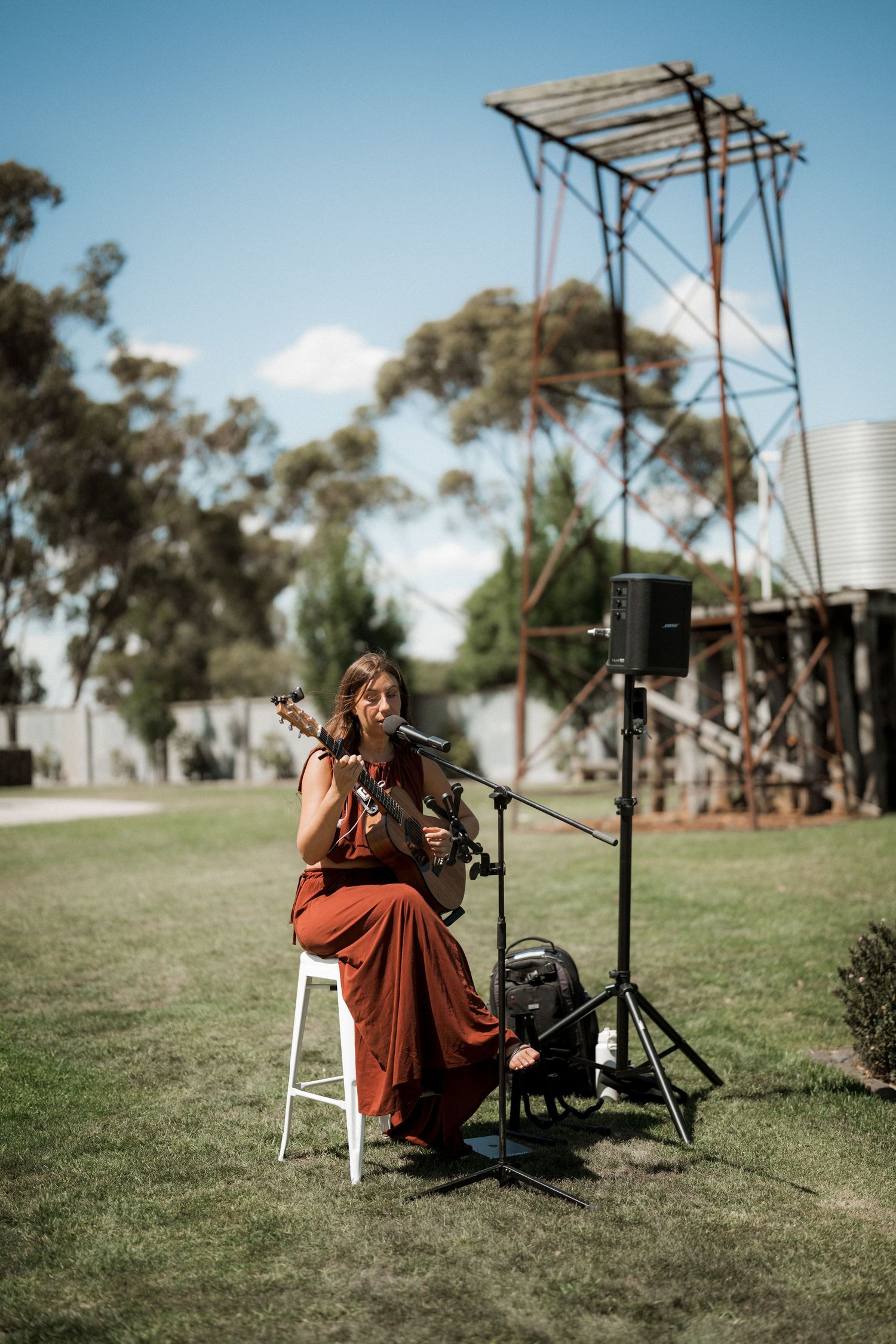 Female musician in rust red dress plays guitar at outdoor wedding ceremony with speaker and microphone setup.