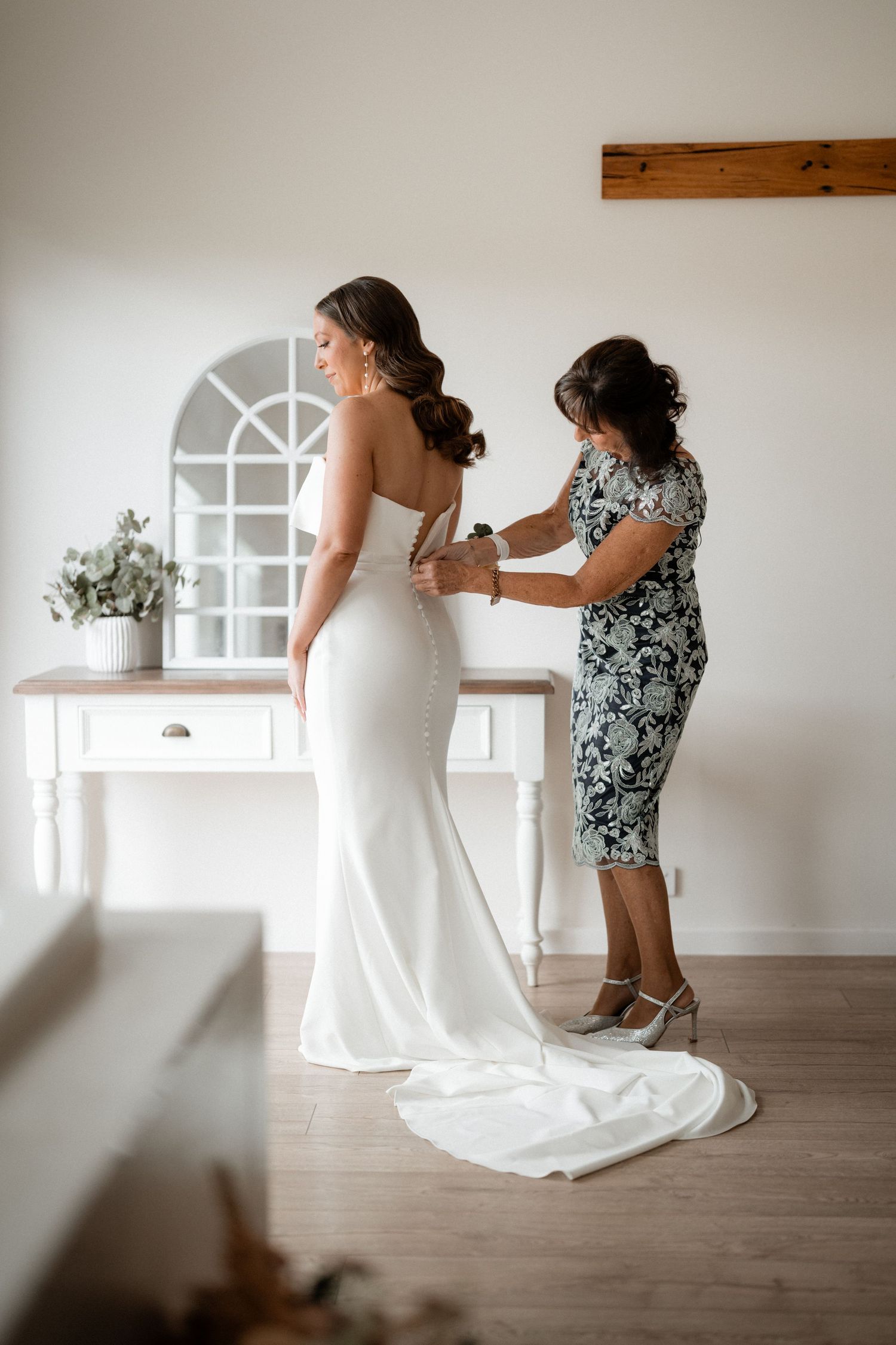 Woman in elegant white wedding dress getting final adjustments in a bright interior room.