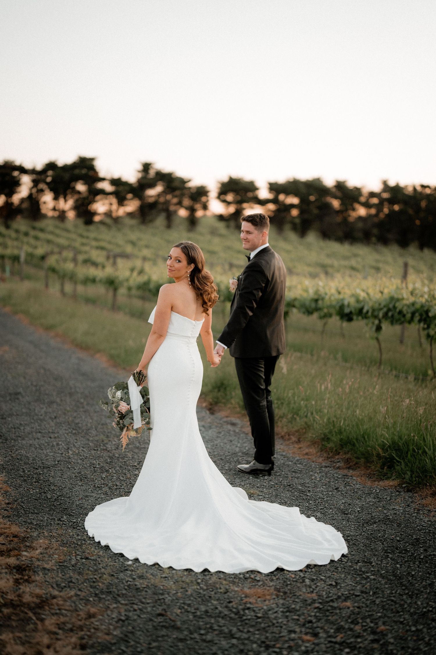 Bride in flowing white dress and groom standing on path by vineyard at golden hour.