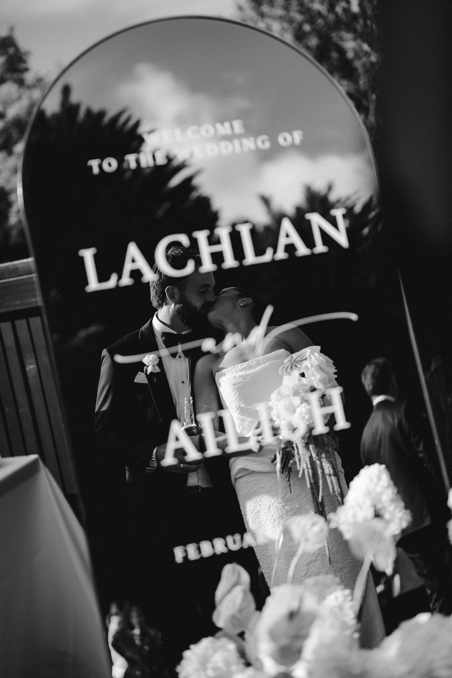 Black and white photo of bride and groom kissing behind wedding welcome sign for Lachlan and Ailish.
