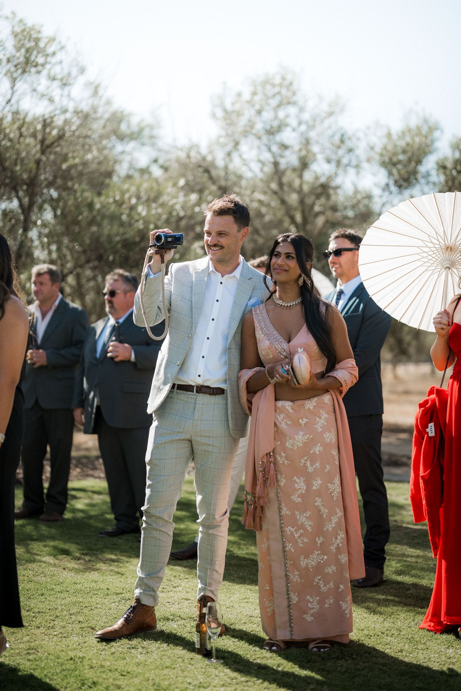 Wedding guests with parasols gathered outdoors under olive trees.