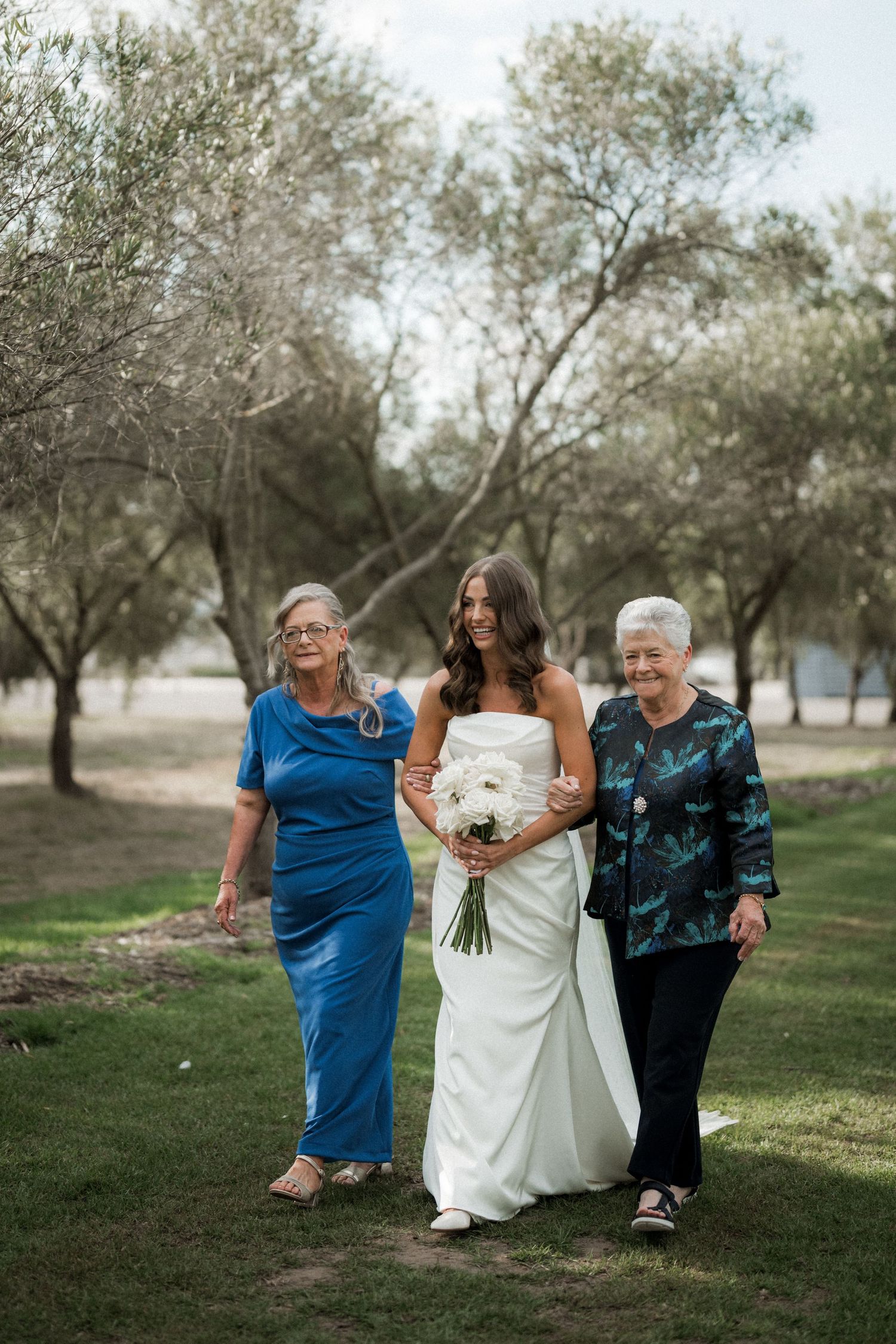 Bride in white dress walks with two older women through an olive grove.