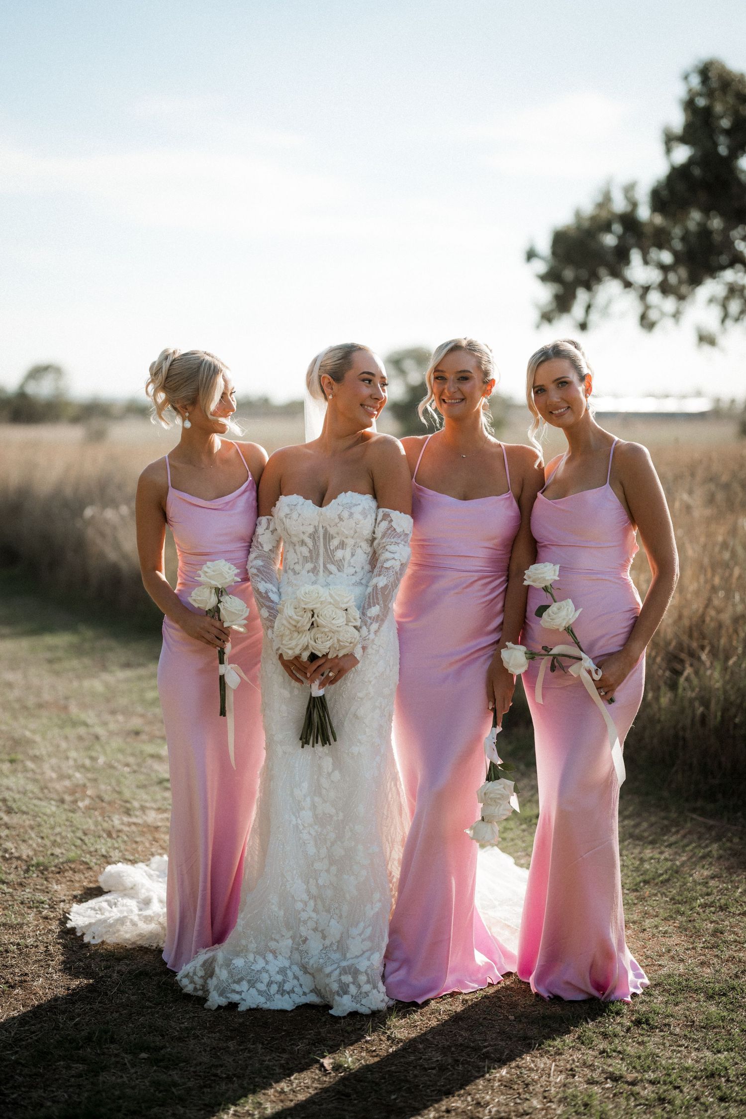 A bride in white lace gown poses with three bridesmaids wearing pink fitted dresses during golden hour.