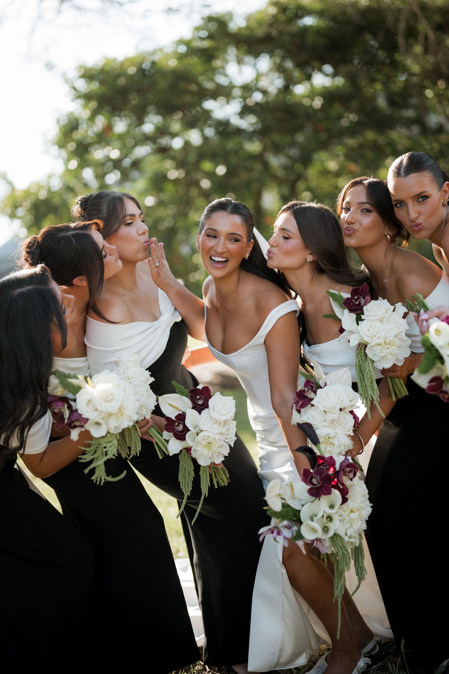 Bridal party in black dresses and white gowns holding bouquets of white and burgundy flowers.