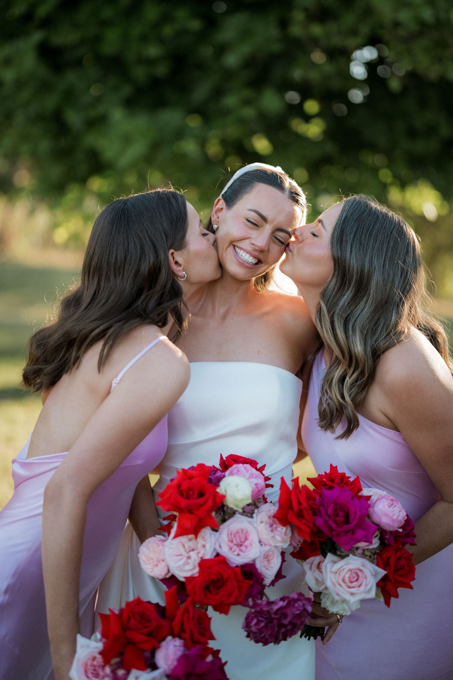 A wedding party in pink dresses poses with vibrant red and pink floral bouquets in an outdoor garden setting.