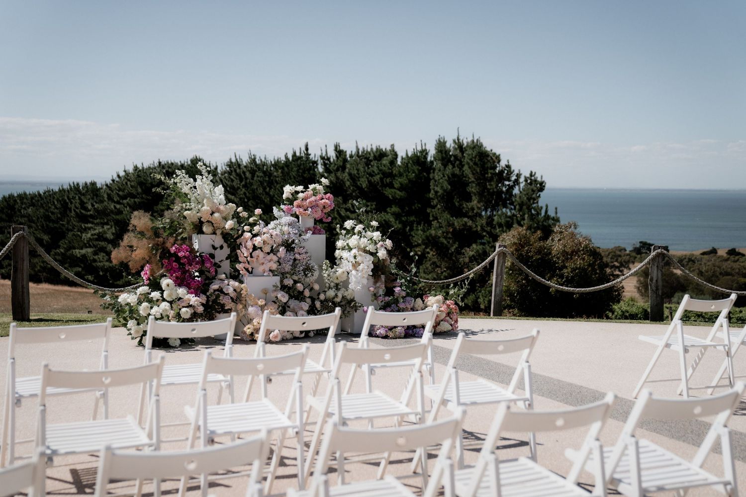 White chairs arranged for an outdoor wedding ceremony with floral arrangements and ocean views in the background.