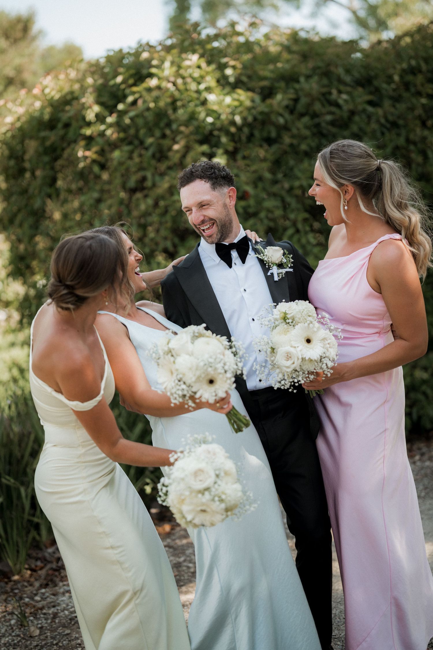Wedding party in formal attire sharing laughs while holding white floral bouquets.