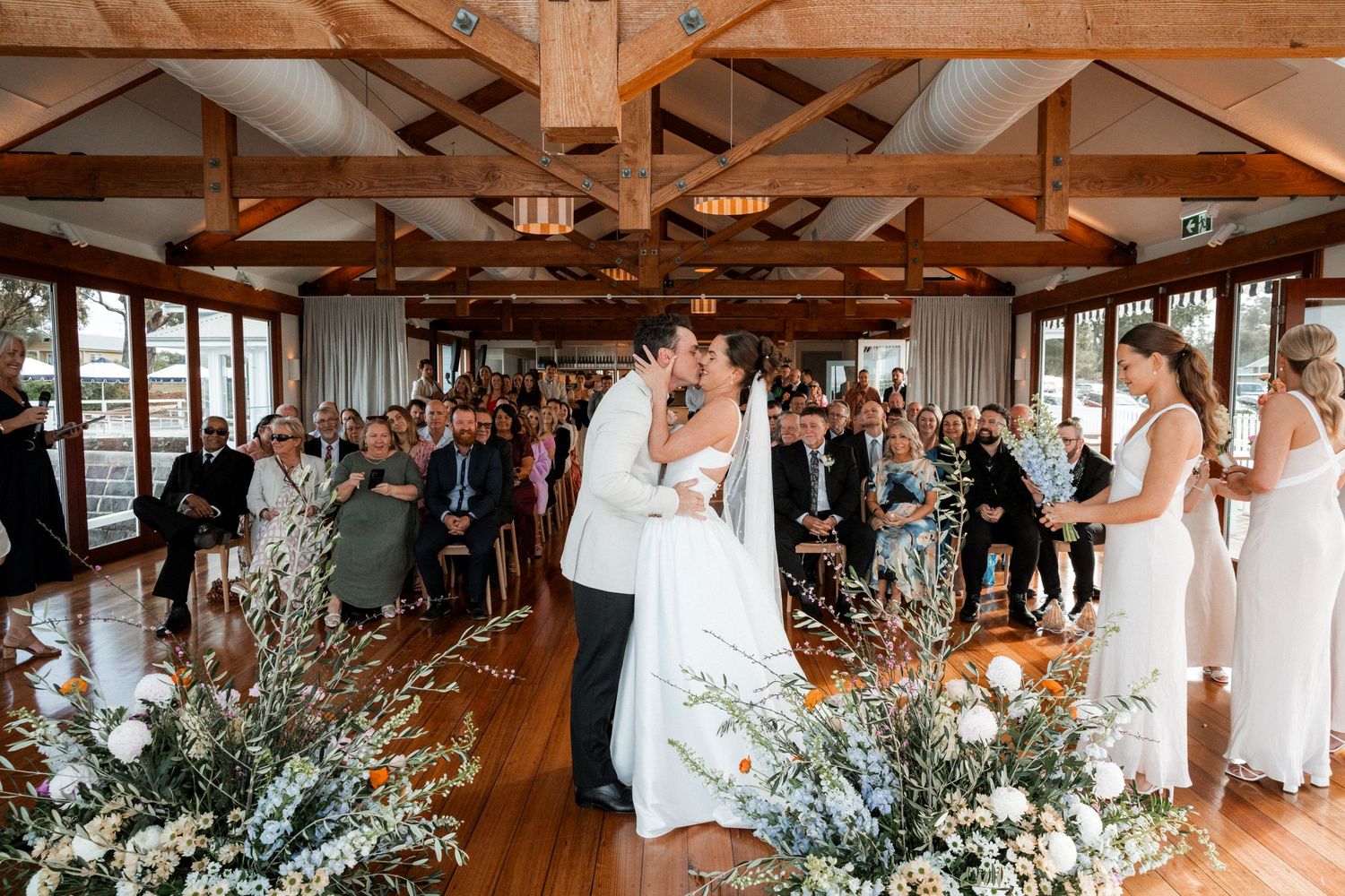 A rustic wooden venue decorated with floral arrangements hosts a wedding celebration with guests in white attire.