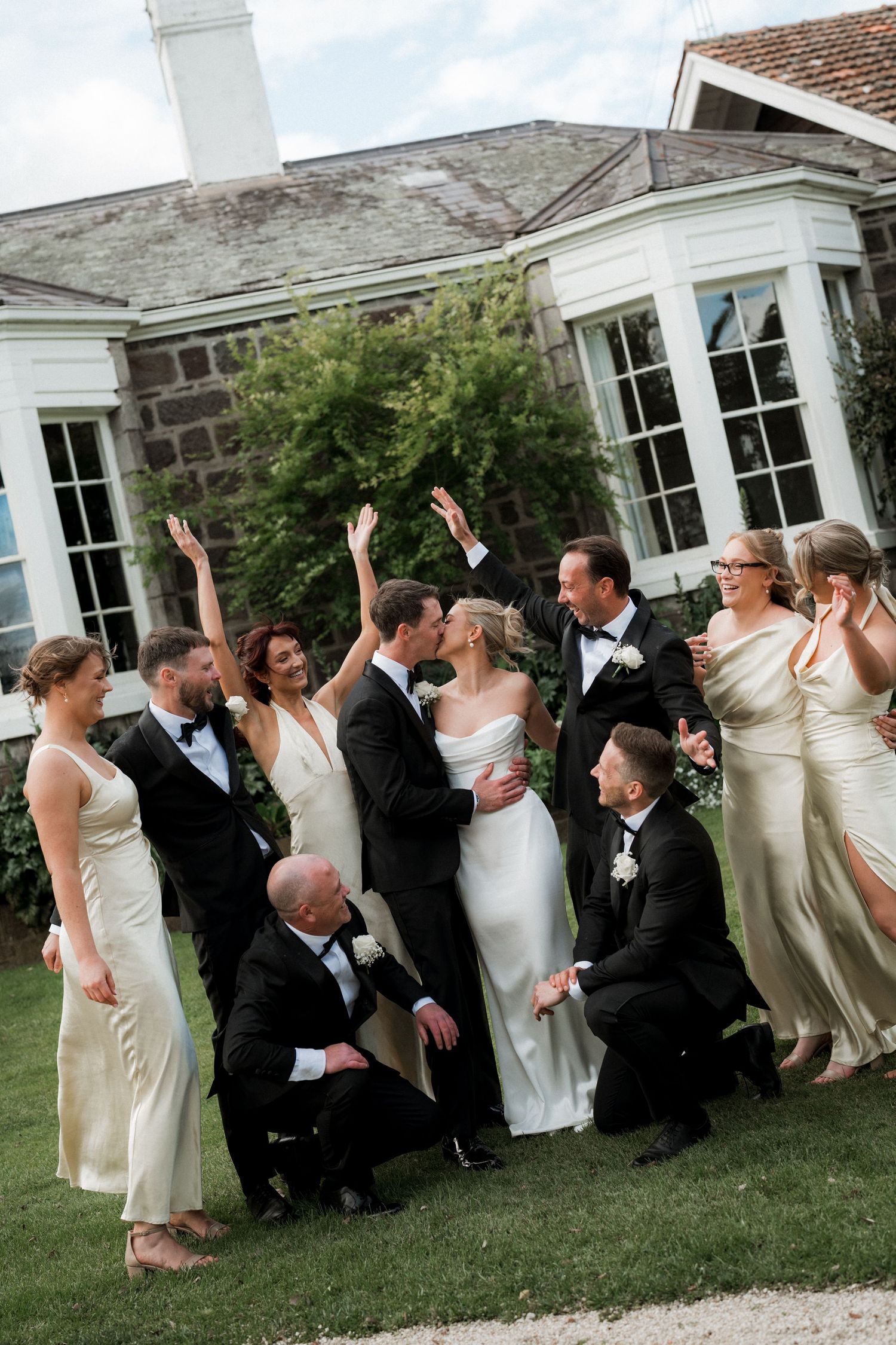 Wedding party in formal attire posing playfully on lawn in front of stone mansion.