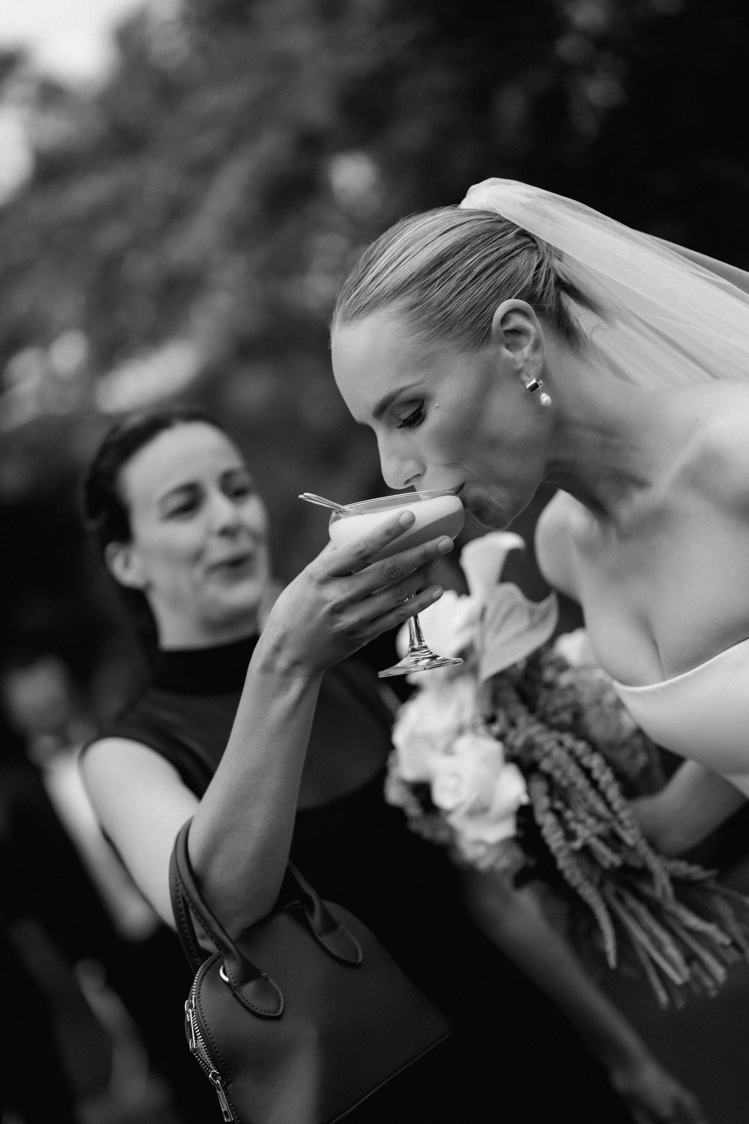 Bride in white dress with veil sips from a glass while holding a floral bouquet.