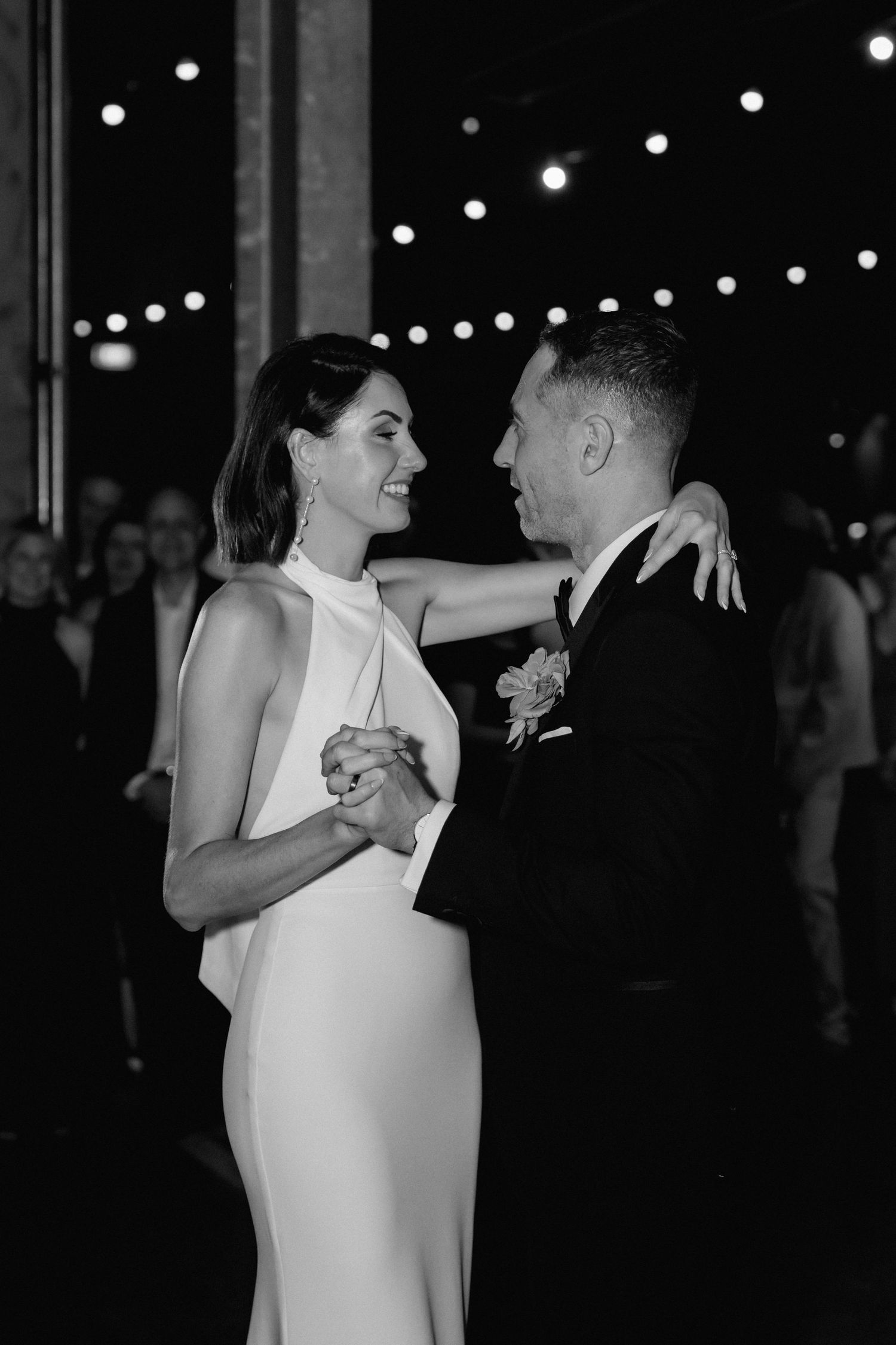 A couple shares an intimate first dance at their wedding reception under twinkling string lights in black and white.