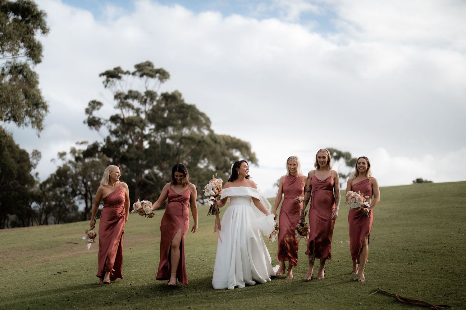Bridal party walks together on grassy hilltop during outdoor wedding ceremony with eucalyptus trees in background.