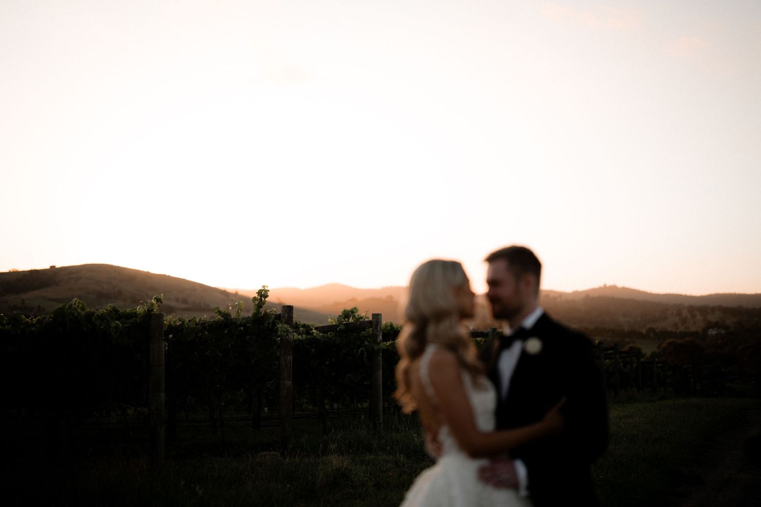 A romantic wedding couple embraces at sunset in front of rolling vineyard hills.