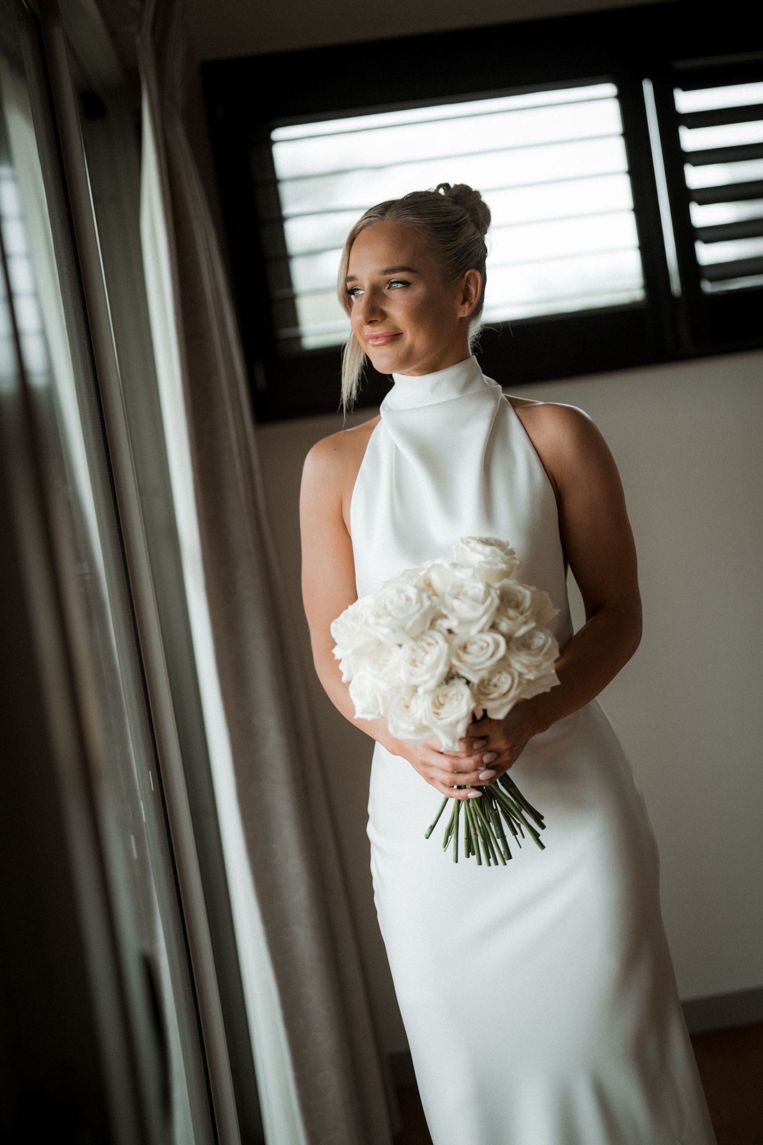 Elegant bride in sleek white halter wedding dress poses by window holding bouquet of white roses.