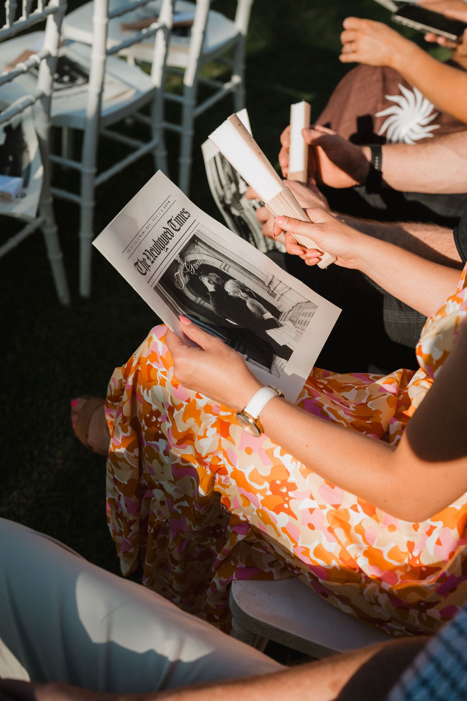 Person in floral dress holds wedding program while seated in outdoor setting.