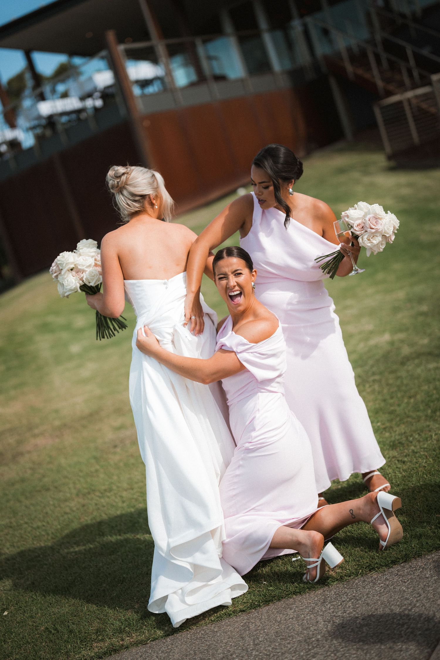 Three bridesmaids in flowing white dresses laugh and dance together while holding bouquets at an outdoor wedding.