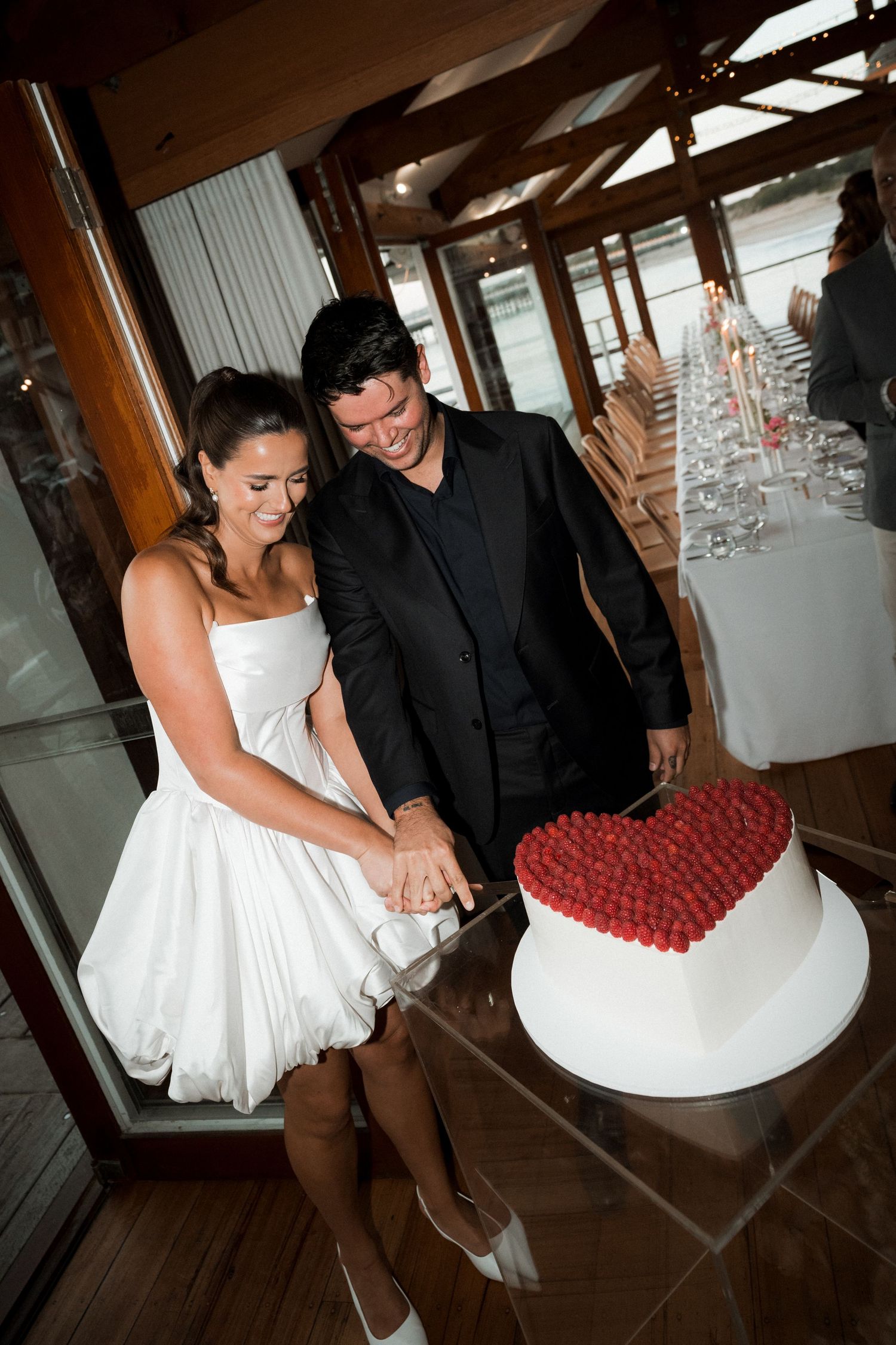 Couple in wedding attire cuts into a white cake topped with red heart-shaped decoration at an indoor reception venue.