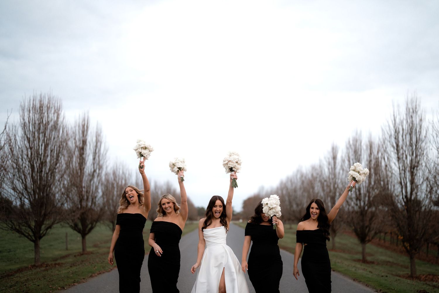 Bridesmaids in black dresses celebrate with the bride in a winter orchard setting.