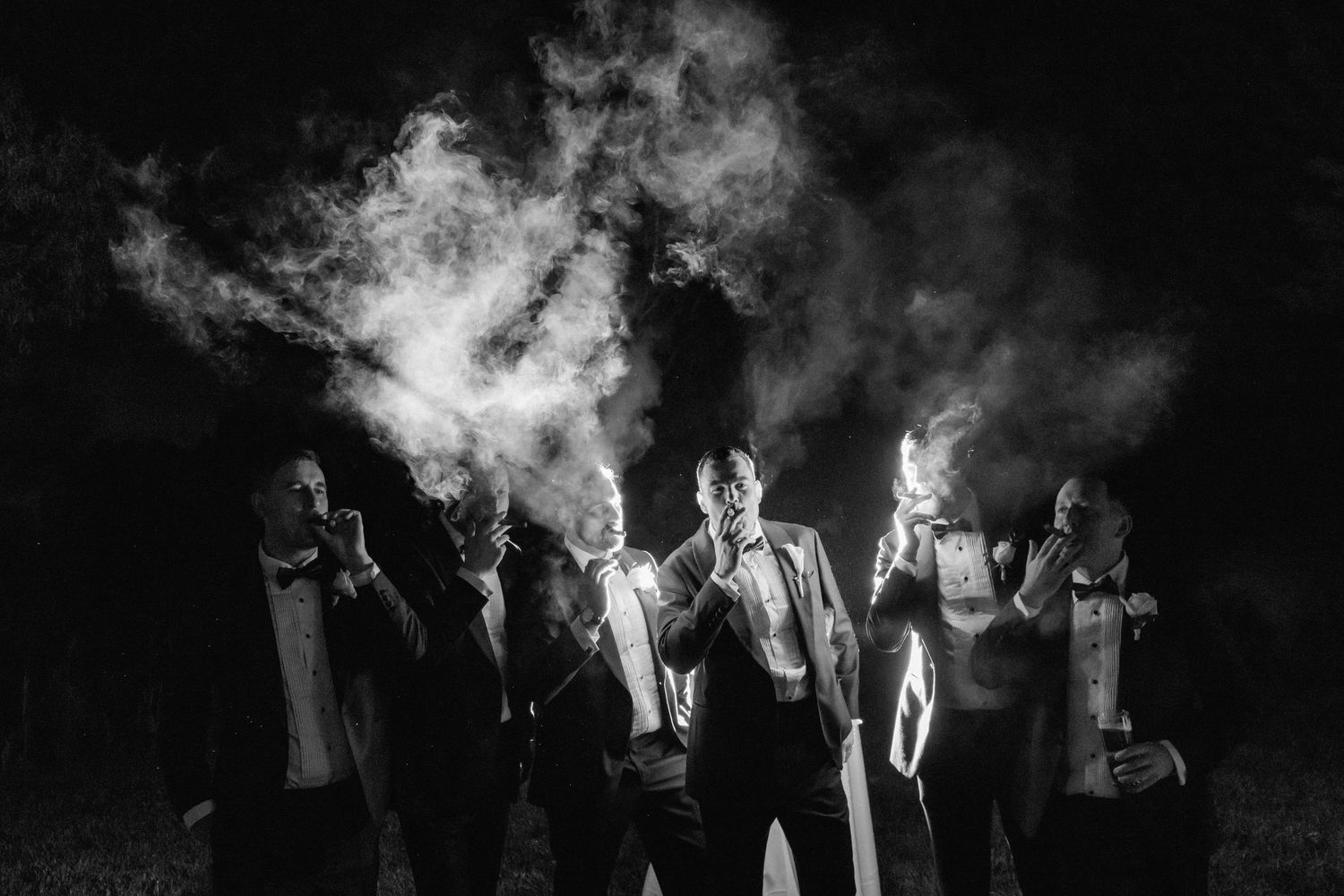 Black and white photo of groomsmen in tuxedos holding sparklers with dramatic smoke at night.