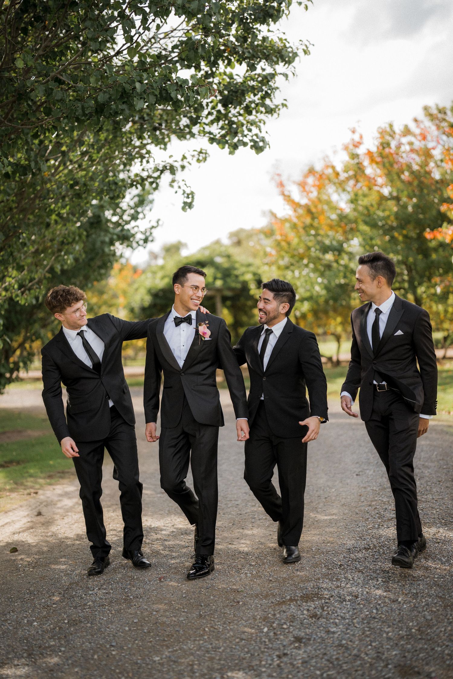 Four groomsmen in black suits walking together on a path surrounded by autumn trees at a wedding.