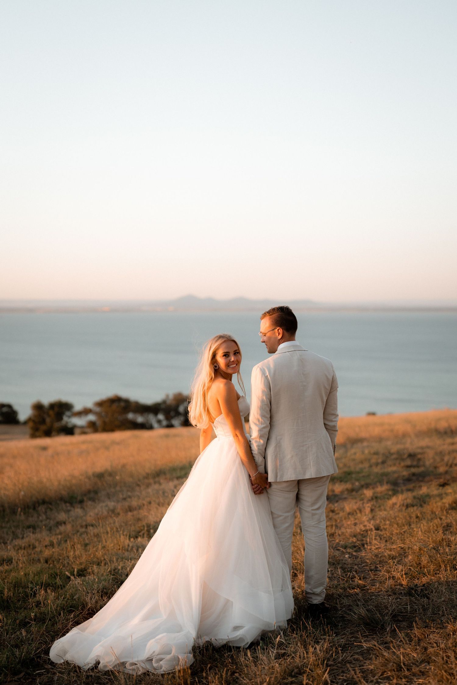 Couple in wedding attire walks hand in hand on hilltop overlooking ocean at sunset.