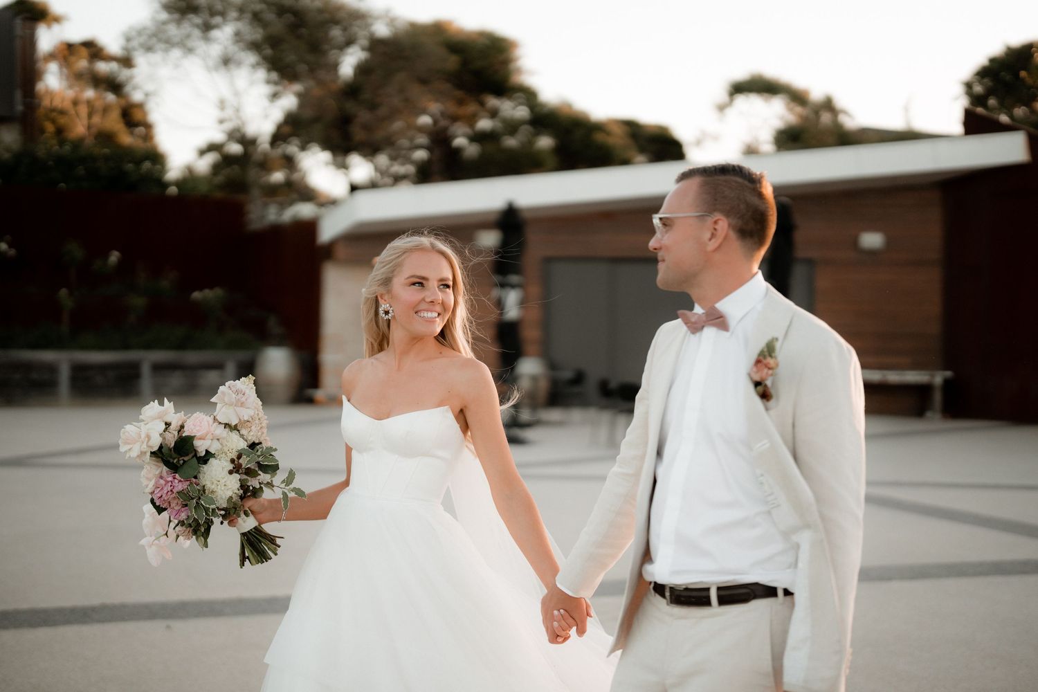Couple in wedding attire walking together outdoors at sunset near brick buildings.