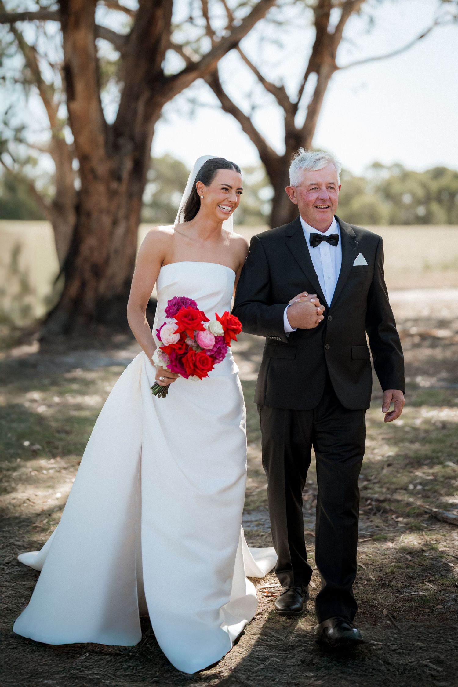 Bride in white dress with red bouquet walking with companion in black tuxedo through outdoor garden setting.