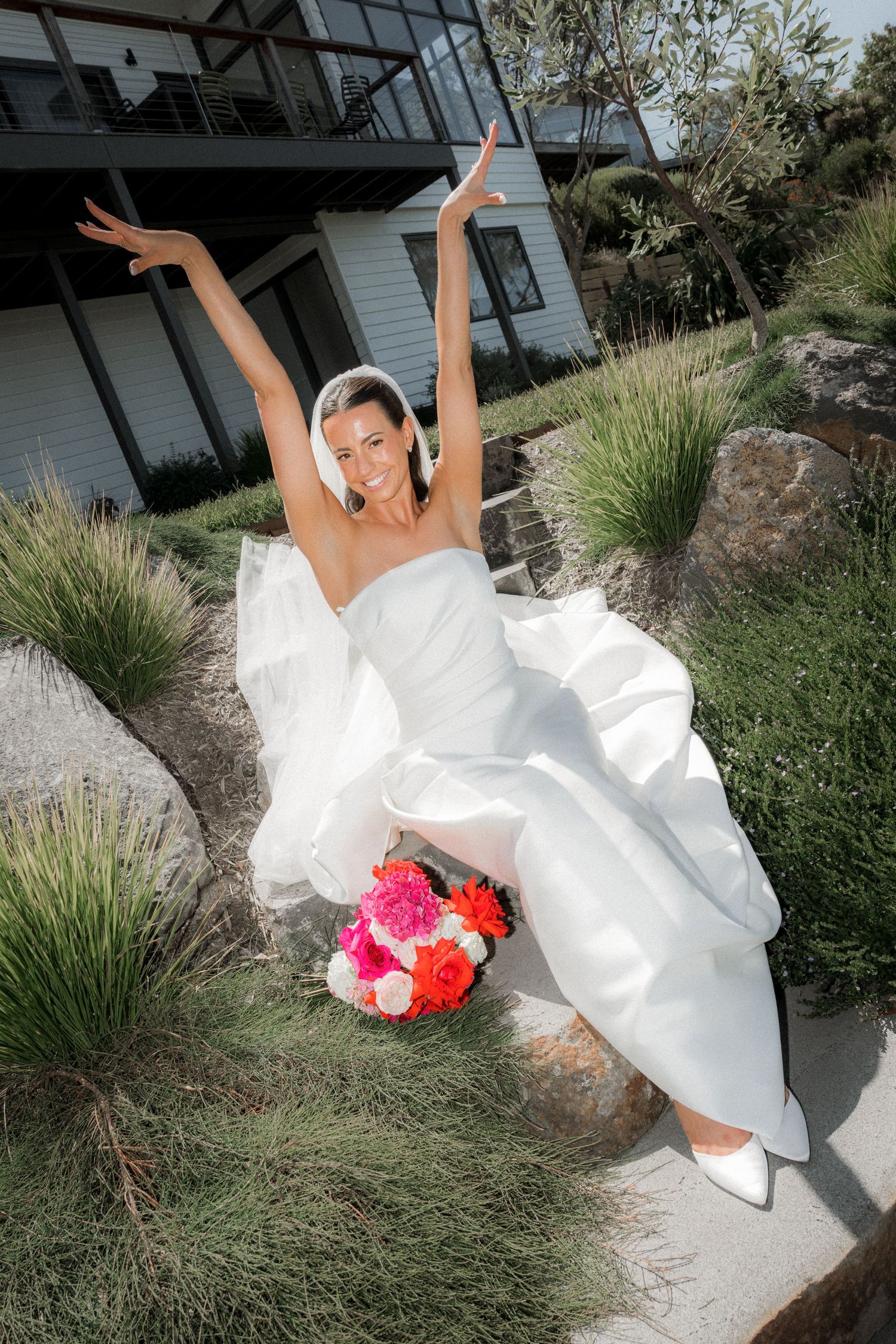 A bride in a flowing white wedding dress poses playfully on landscaped grounds with bright pink and coral bouquet.