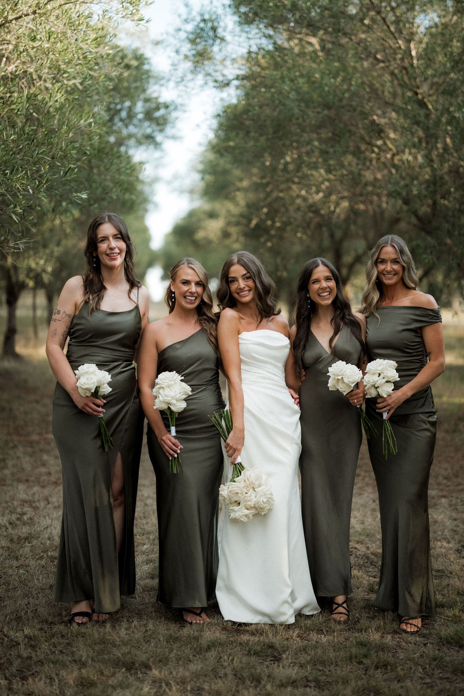 A bridal party in olive green dresses and white wedding gown pose together in an outdoor olive grove setting.