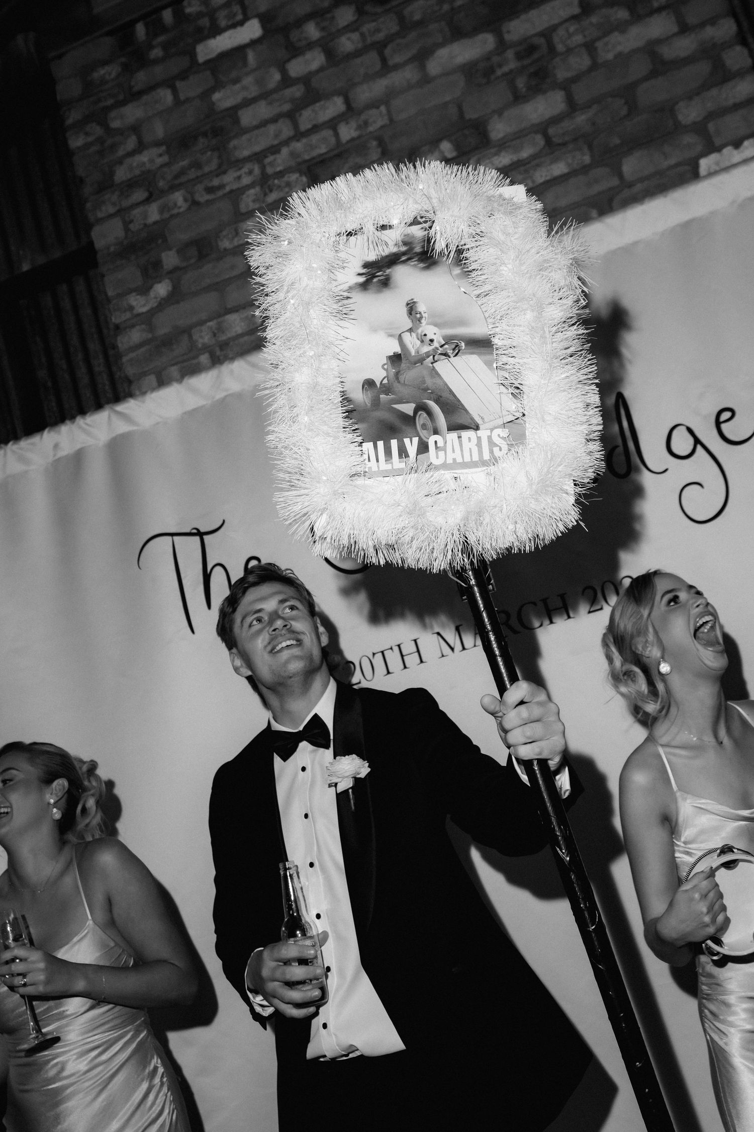 A black and white photo of wedding guests celebrating with a decorative light-up party sign.