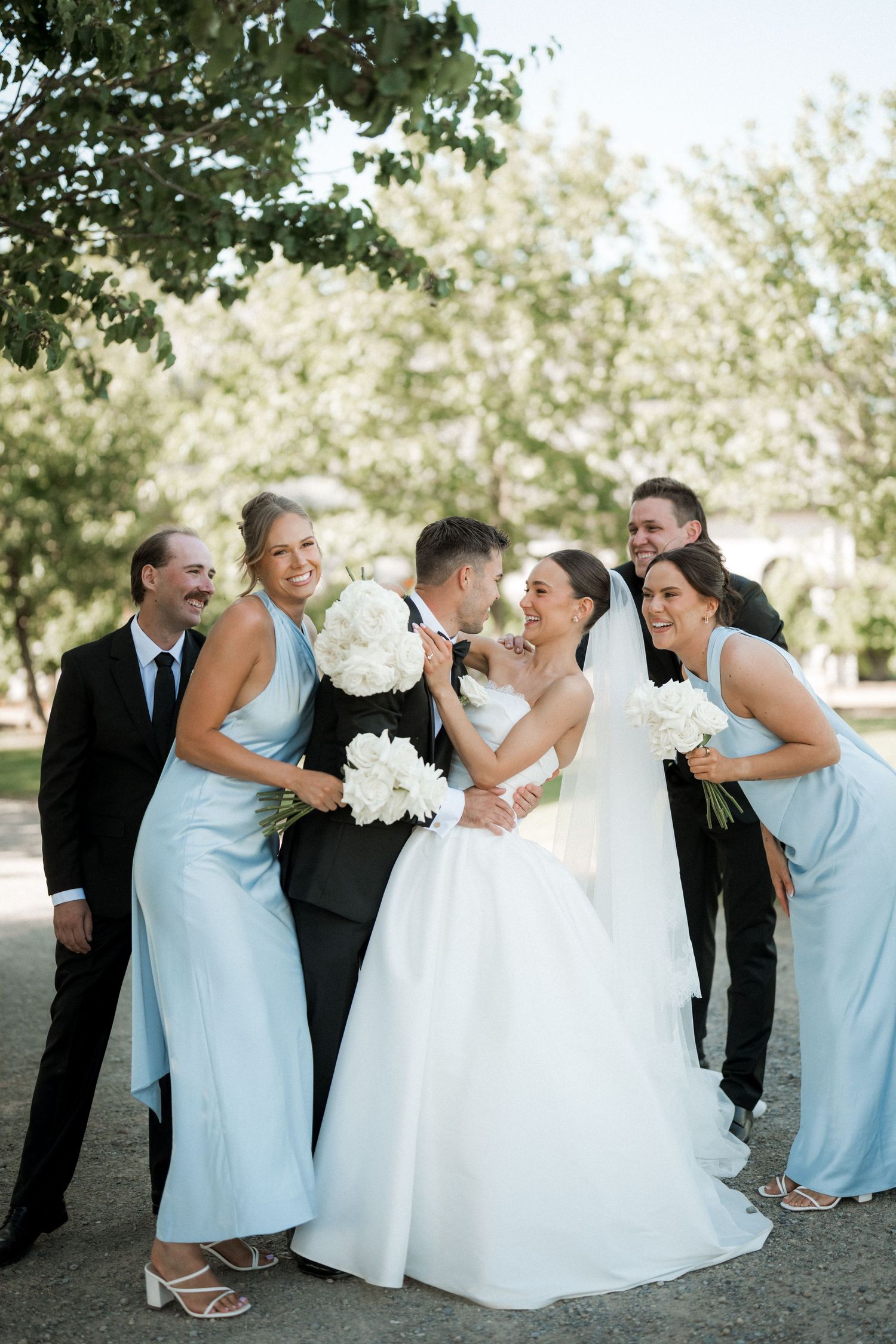 Bridal party in blue dresses and white gown posing playfully under trees.