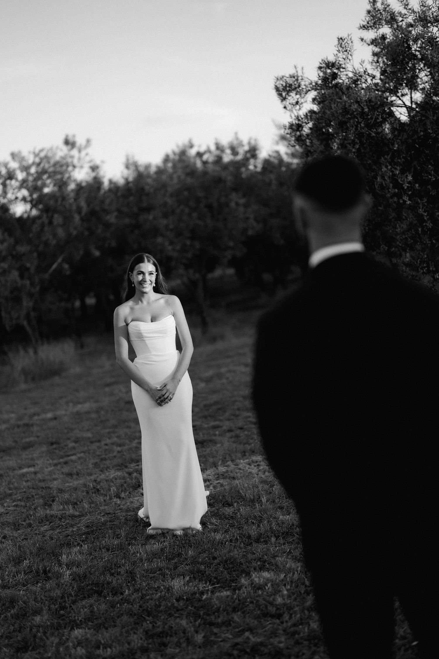 A bride stands in a white dress against a backdrop of trees in black and white photography.